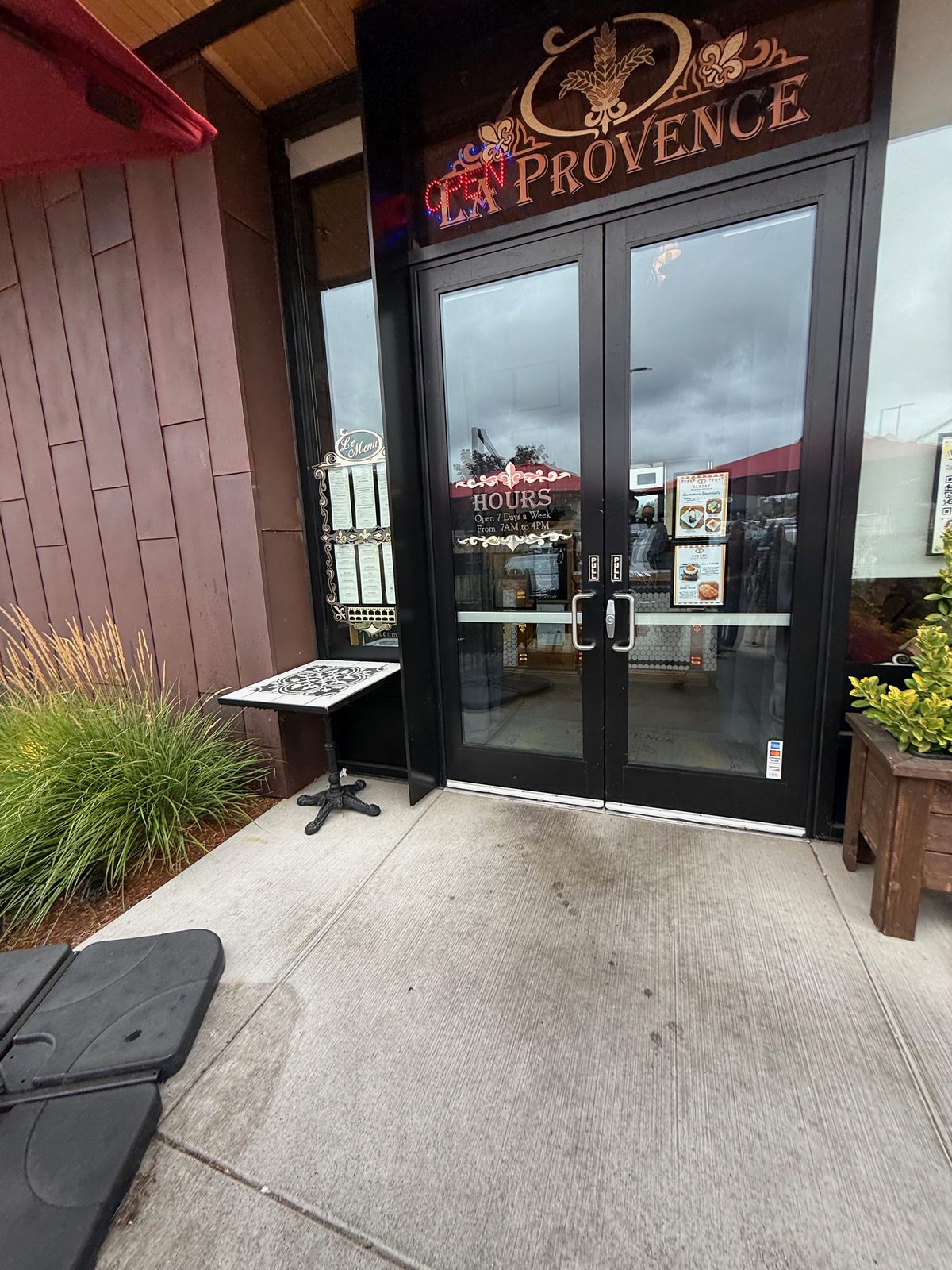 The black double doors of La Provence, with a gold script sign above reading La Provence and a red neon OPEN sign, a menu in a wrought-iron stand to the left, and a window card reading HOURS, Open 7 Days a Week, From 7AM to 4PM