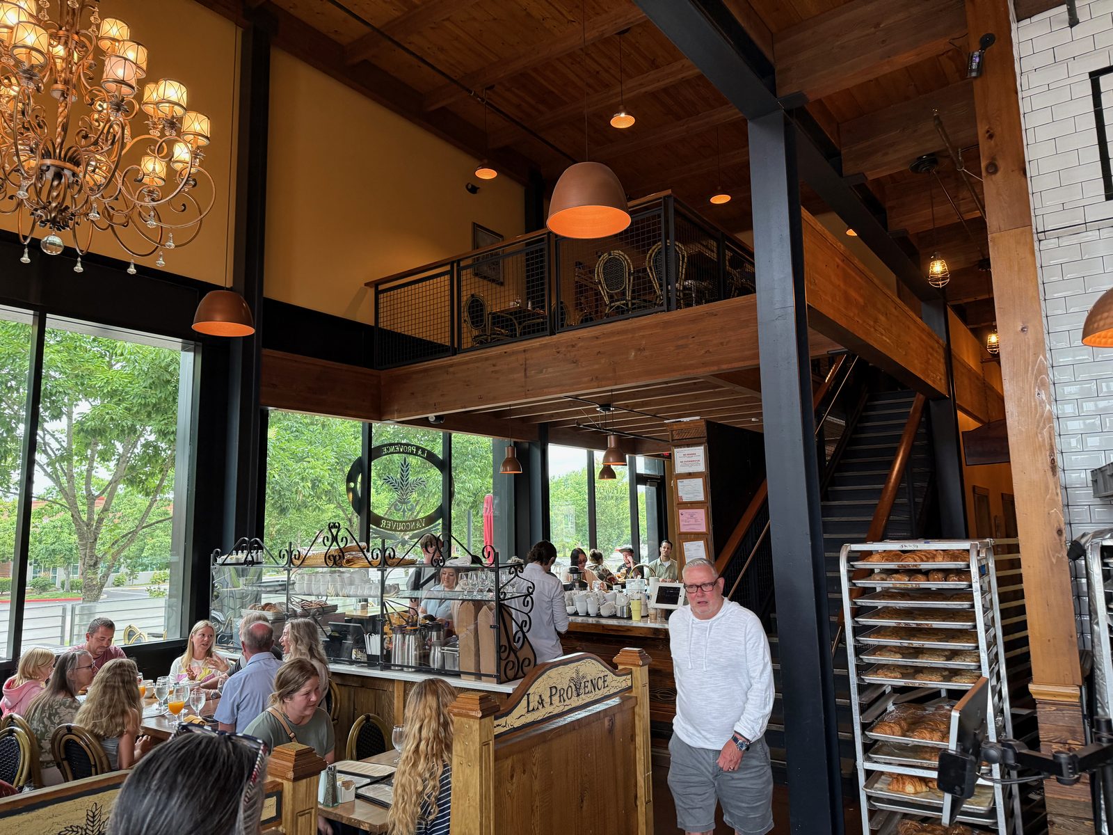 The interior of La Provence looking toward the front, with a high timber ceiling, a mezzanine level of bistro tables behind a black railing, copper pendant lamps, a full dining room below, a circular window sign reading La Provence Vancouver, and tall racks of croissants to the right