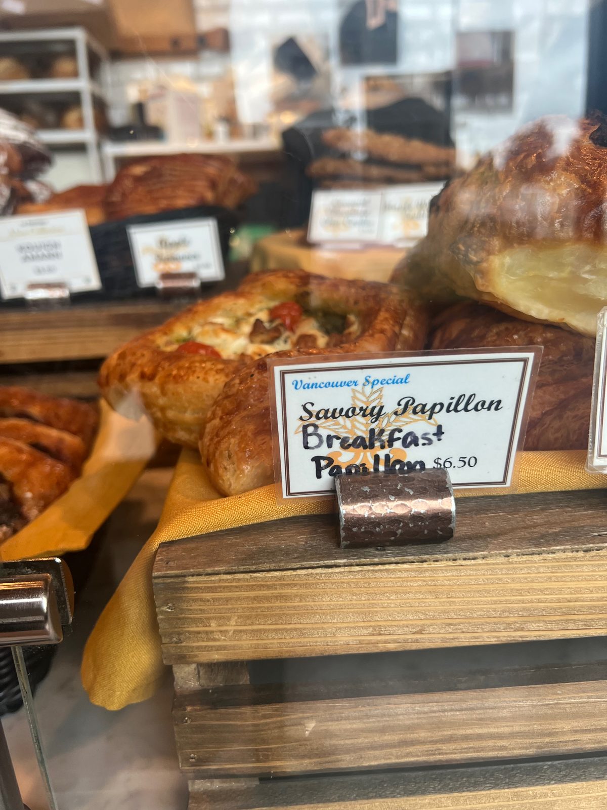 A close view of a handwritten card in the La Provence pastry case reading Vancouver Special in blue ink above Savory Papillon Breakfast Papillon, 6.50, propped in a metal holder in front of a flat golden pastry topped with cherry tomatoes, herbs and cheese