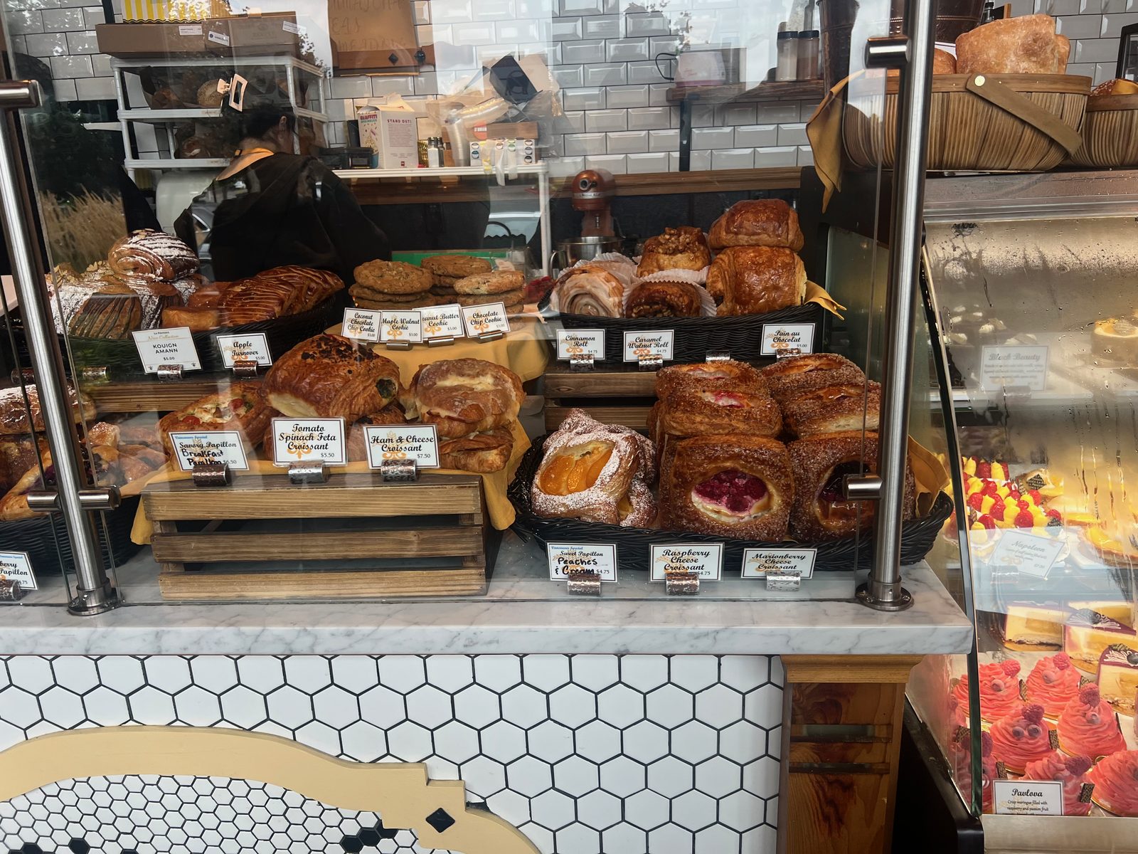 The pastry case at La Provence seen through glass, holding kouign-amann, apple turnovers, savory papillons topped with tomato and herbs, ham and cheese croissants, sugar-dusted peaches and cream pastries, raspberry and marionberry cheese croissants, cookies, cinnamon and caramel walnut rolls and pain au chocolat, each with a small handwritten card