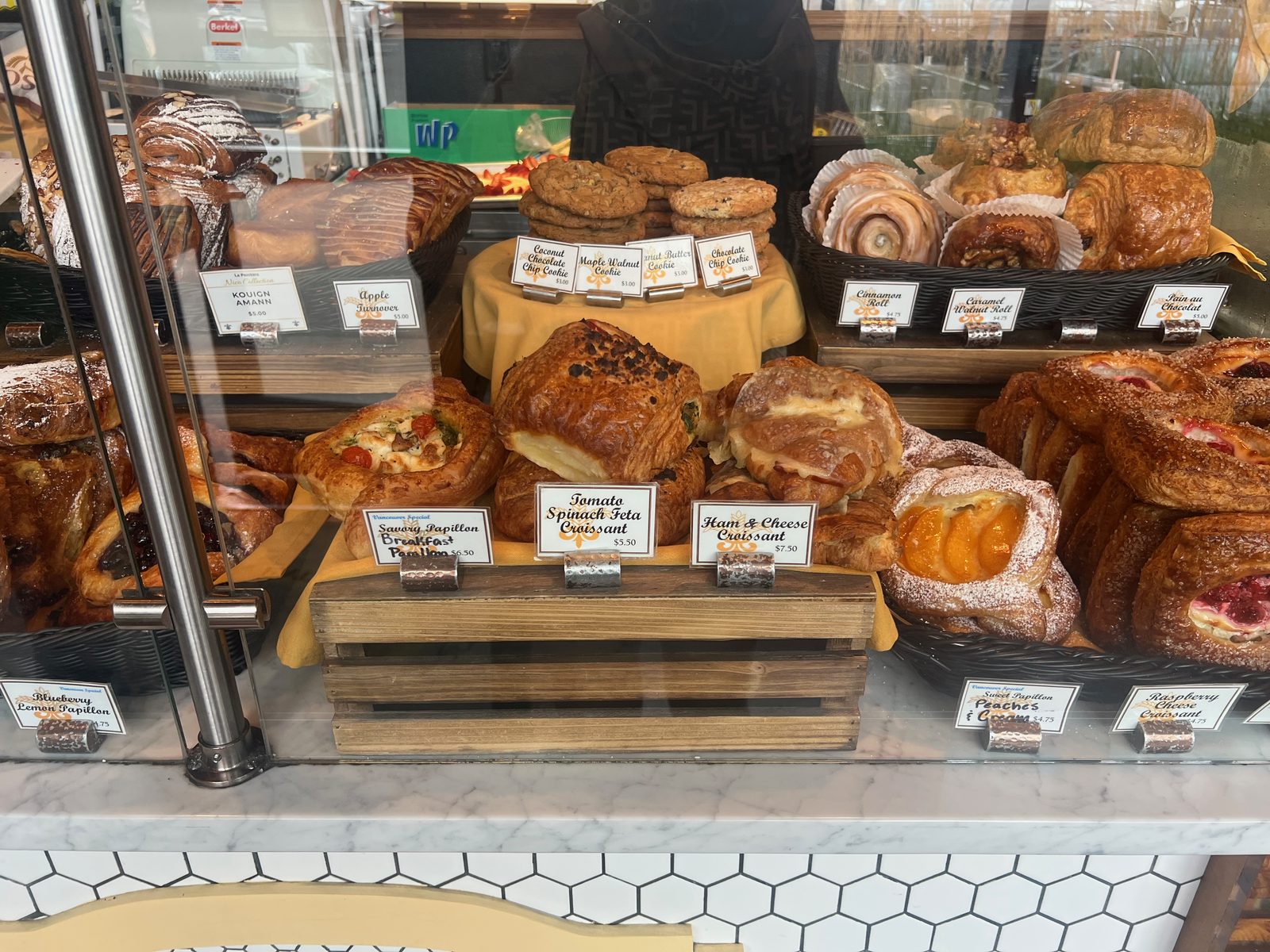 A closer view of the La Provence pastry case with handwritten price cards reading Kouign Amann 5.00, Apple Turnover, Savory Papillon Breakfast Papillon 6.50, Tomato Spinach Feta Croissant 5.50, Ham and Cheese Croissant 7.50, Blueberry Lemon Papillon 4.75, Cinnamon Roll 4.75, Caramel Walnut Roll, Pain au Chocolat and cookies
