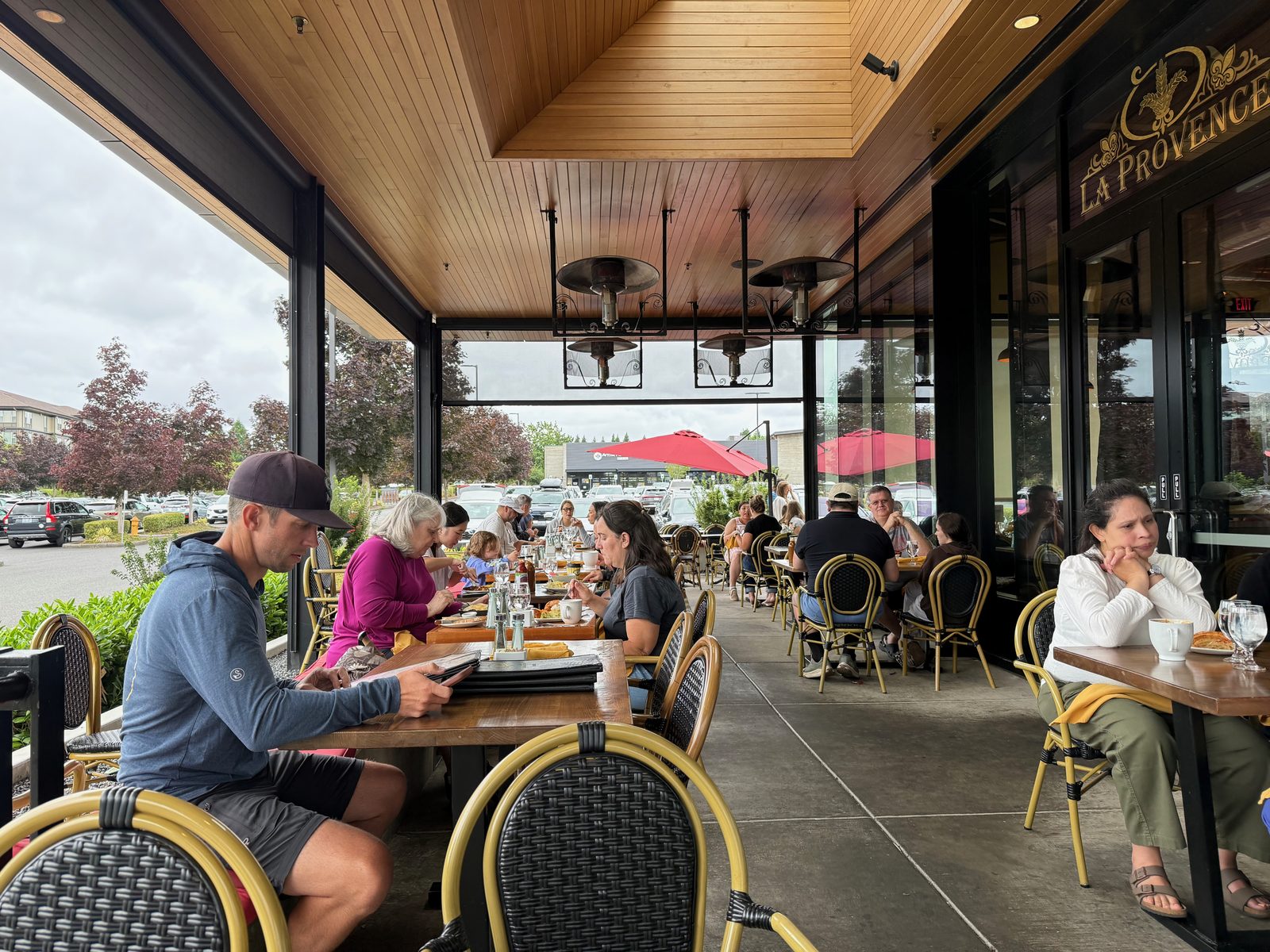 The covered patio at La Provence, a long run of yellow-framed bistro chairs and wooden tables under a warm timber ceiling with overhead heaters, every table occupied, the parking lot and red maples visible through the open side