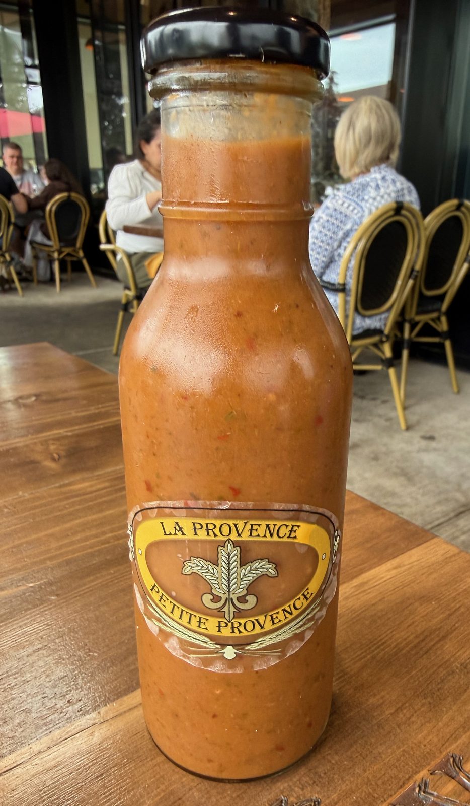A glass bottle of La Provence house sauce on a wooden table, filled with a thick orange-red sauce visibly flecked with pepper and seeds, with a black lid and a gold and yellow oval label reading LA PROVENCE above and PETITE PROVENCE below, a wheat sheaf and fleur-de-lis between them