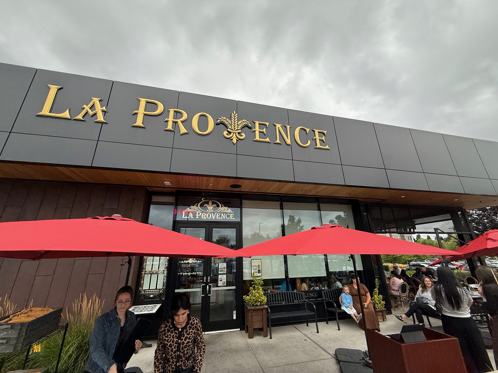 The front of La Provence in Vancouver, Washington on an overcast Saturday morning — a dark grey façade with LA PROVENCE in tall gold serif capitals, a gold wheat sheaf and fleur-de-lis standing in for the V, red patio umbrellas open below, and every outdoor table occupied