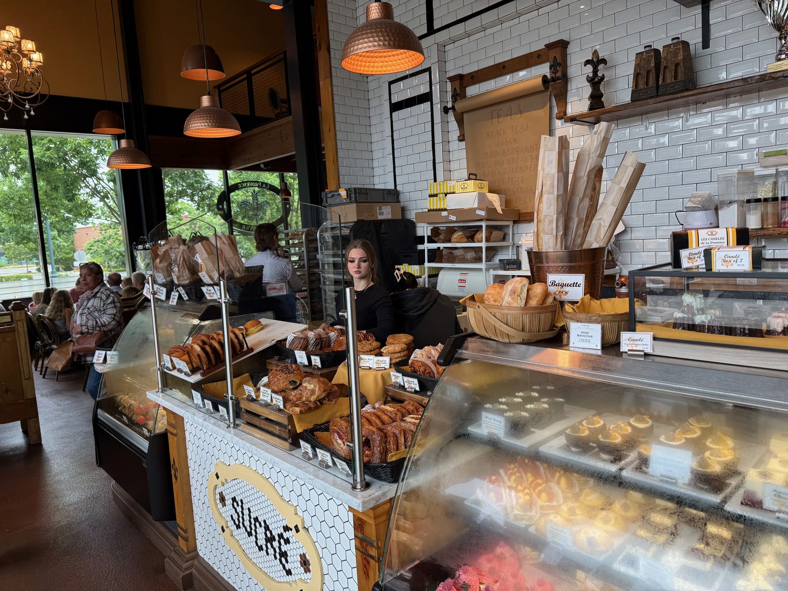 The long pastry counter at La Provence with SUCRÉ set in black tile on white mosaic along the front, glass cases of tarts, éclairs and fruit pastries, a copper bucket of baguettes, a staff member working behind the case, and diners at tables by the window