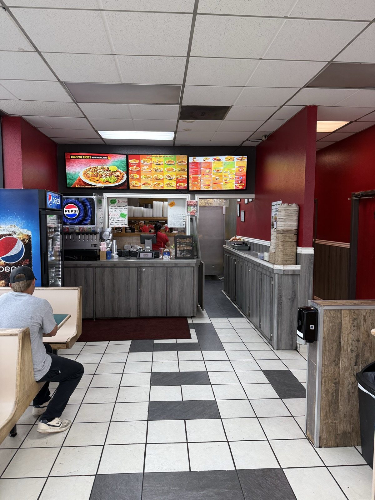 The dining room and order counter at Muchas Gracias Mexican Food — deep red walls, a black-and-white checkerboard tile floor, three lit digital menu screens overhead with one advertising birria fries, a Pepsi fountain and cooler at the left, a handwritten New Hours notice taped in the service window, staff in red polo shirts working behind the glass, a doorway marked PICK-UP HERE, and one customer eating alone in a booth