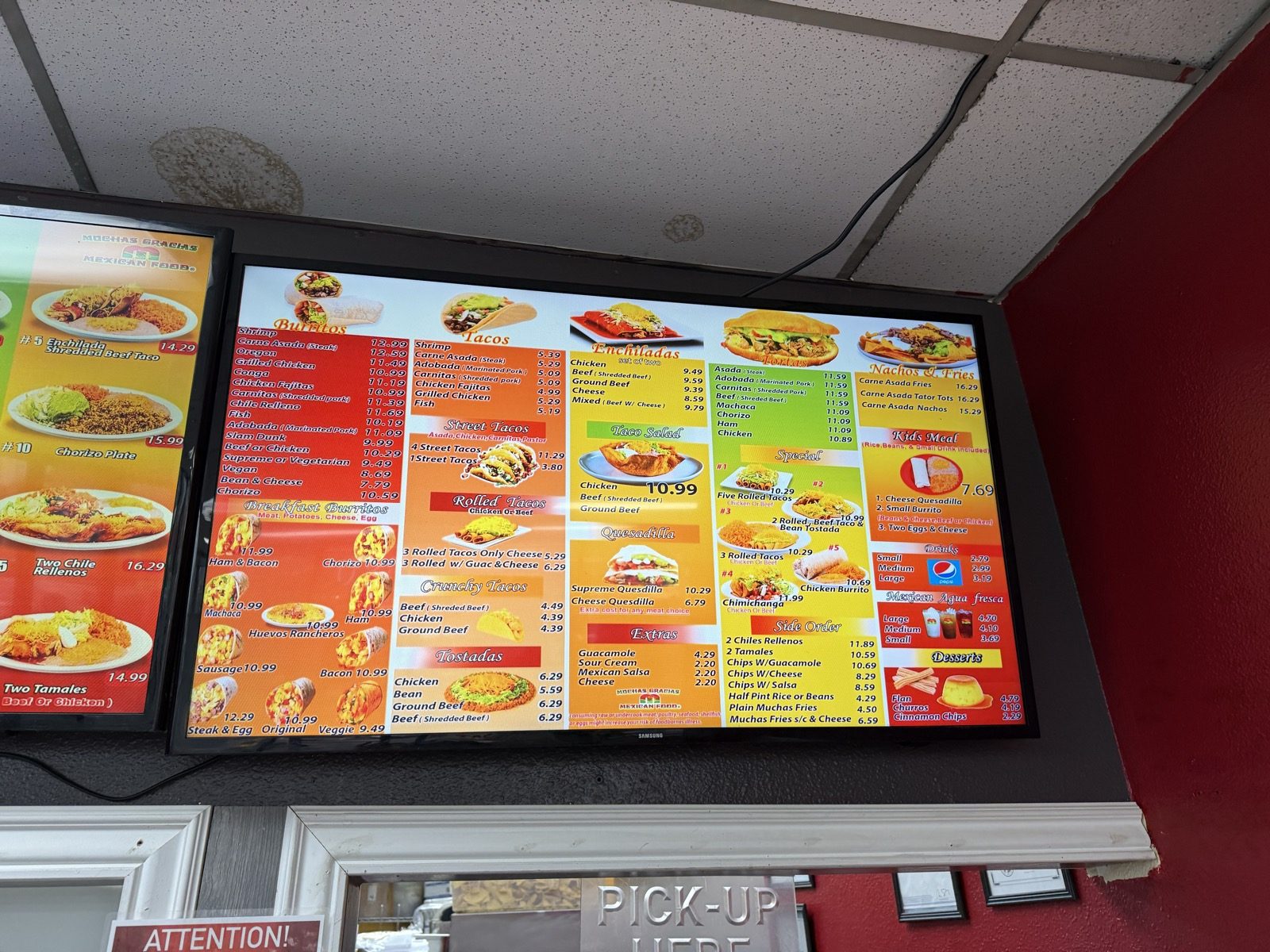 A second digital menu screen listing burritos, tacos, street tacos, rolled tacos, enchiladas, tostadas, quesadillas, tortas, nachos and fries, kids meals, drinks and desserts with prices, mounted above a doorway marked PICK-UP HERE on a red wall