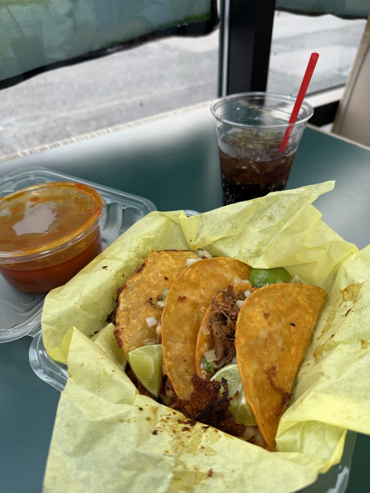 Four quesabirria tacos in a clear clamshell on yellow paper, corn tortillas fried orange in birria fat and filled with shredded beef, white onion and cilantro with lime wedges, next to a clear cup of thin orange consomé and a cola with a red straw