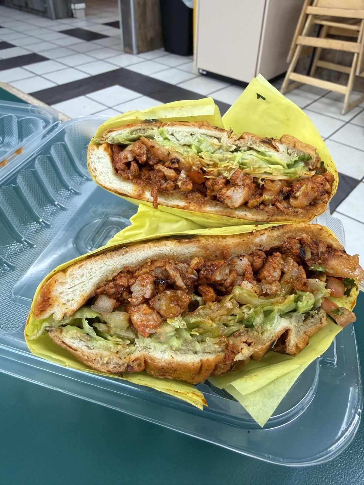 A torta cut in half on yellow paper in a takeout clamshell, both halves face up, a bolillo roll filled with chopped red-marinated griddled meat, shredded lettuce, avocado spread and pico de gallo, on a table above a black-and-white checkerboard tile floor