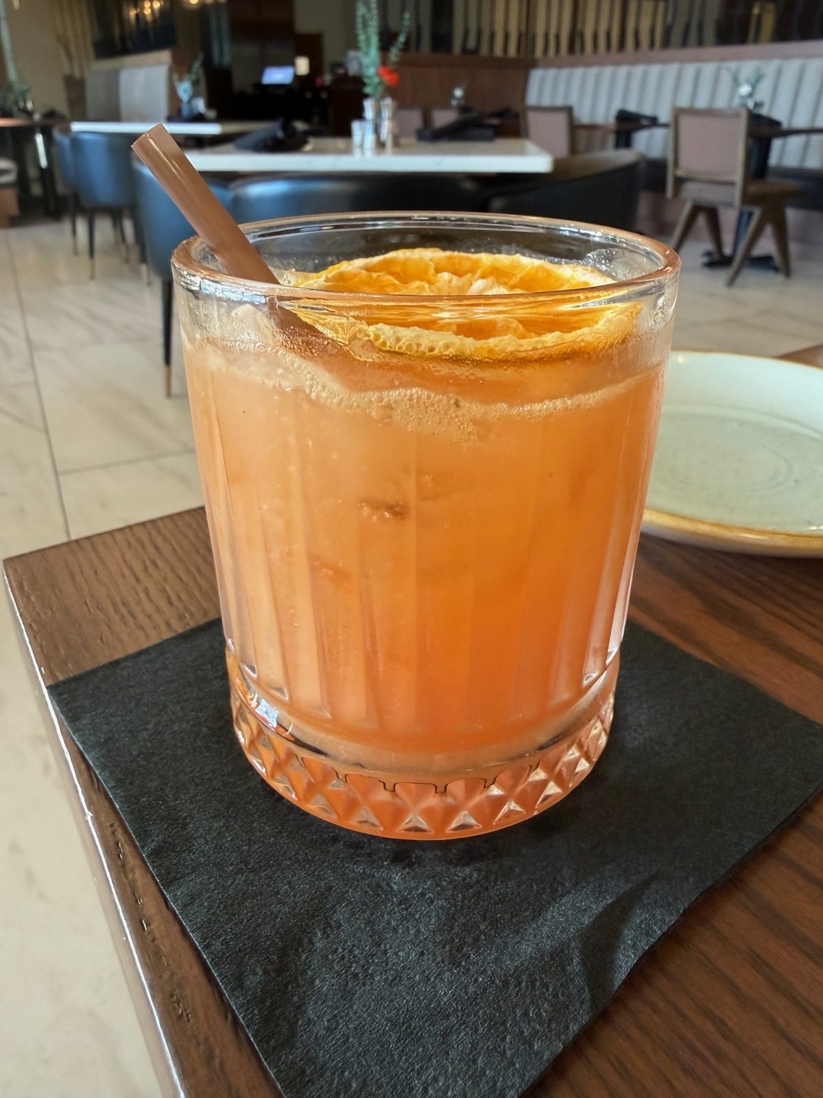 A POG Rum cocktail, orange-colored passionfruit-orange-guava juice and rum in a cut-crystal rocks glass, garnished with a dehydrated orange wheel and served with a metal straw on a black napkin