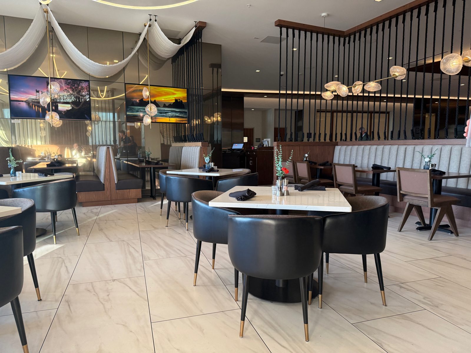 The empty early-evening dining room at Ondus on the Waterfront, marble-pattern tile floor, black barrel chairs at white marble tables, and two backlit sunset photographs on a mirrored wall