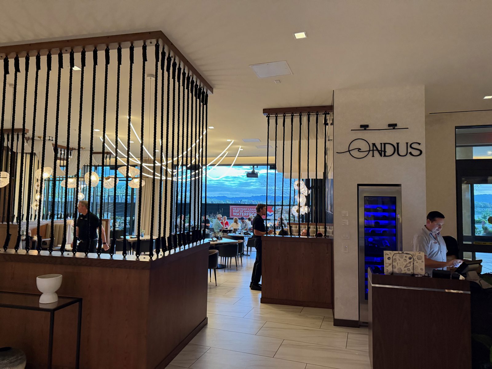 The ONDUS wall sign near the host stand at Ondus on the Waterfront, its logo a simple stylized wave above the wordmark, with a blue-lit glass wine refrigerator beside it and a host checking a tablet at the podium