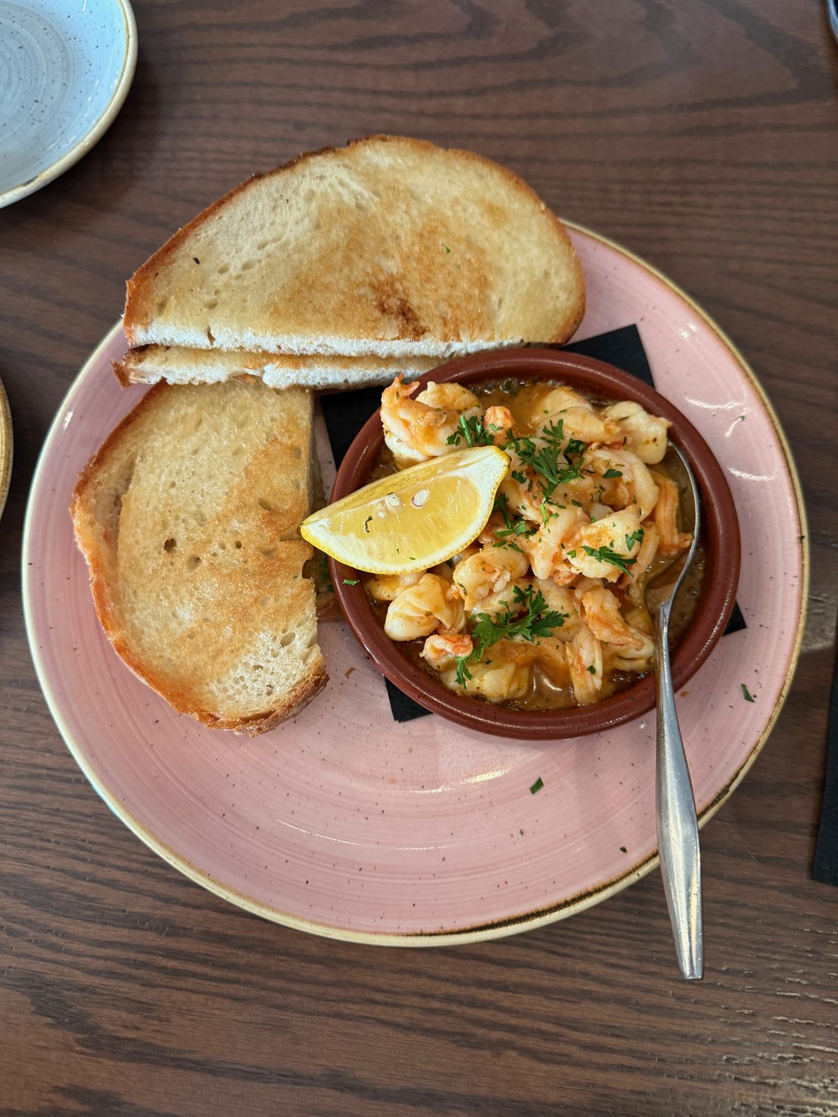 A cazuela dish of sizzling garlic shrimp in a butter sauce, garnished with parsley and a lemon wedge, served on a pink plate with two thick slices of toasted sourdough bread