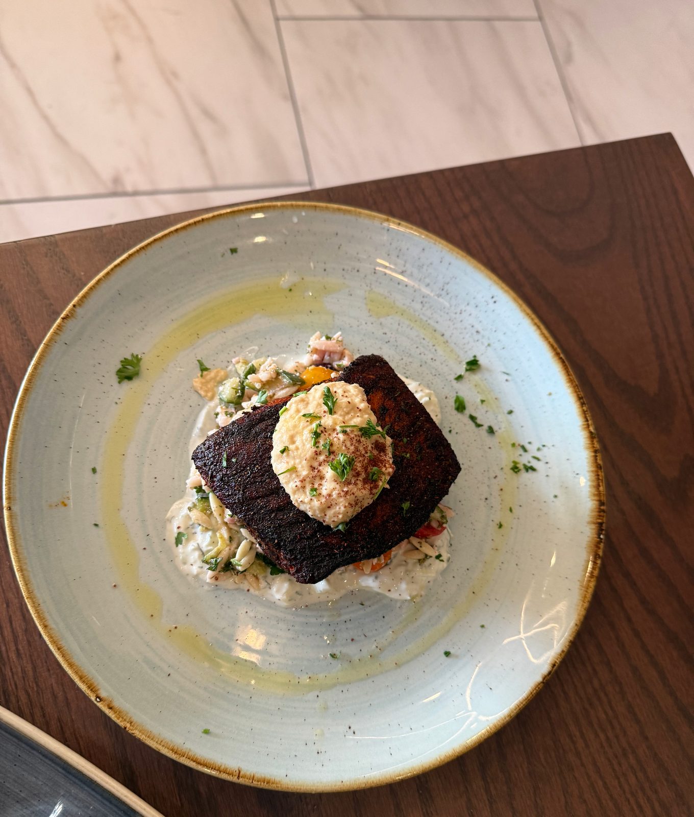 A blackened Mediterranean salmon fillet topped with a dollop of hummus and sumac, served over a creamy orzo salad with tomato and cucumber, drizzled with olive oil