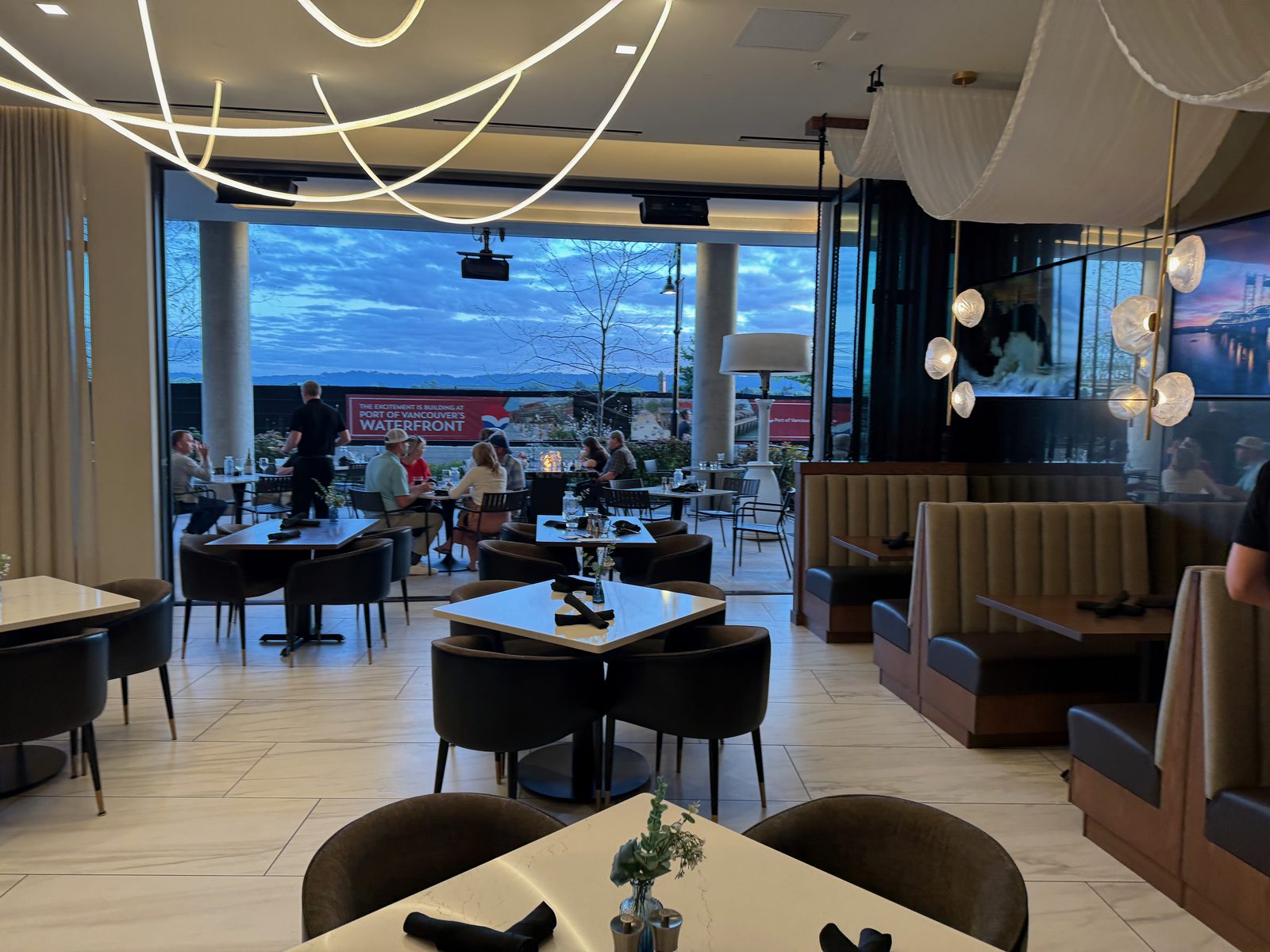 The dining room at Ondus on the Waterfront in Vancouver, Washington, open to a patio at dusk, diners seated both inside and out, with a darkening blue sky over the Columbia River in the distance