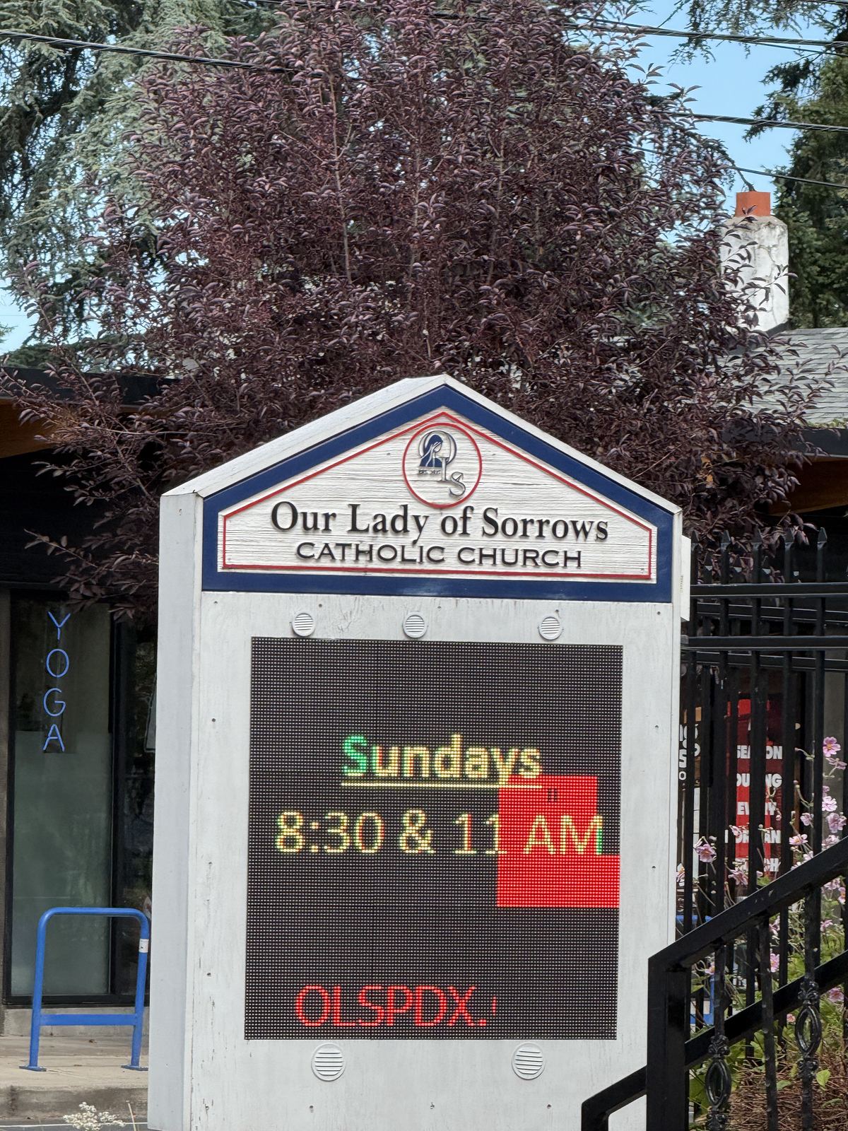 A white peaked-roof church sign with a navy and red border reading "Our Lady of Sorrows — Catholic Church" above a full-colour LED marquee lit with "Sundays 8:30 & 11 AM" and "OLSPDX.", a purple-leaf plum tree behind it, a black wrought-iron fence to the right and the blue window lettering of a yoga studio to the left