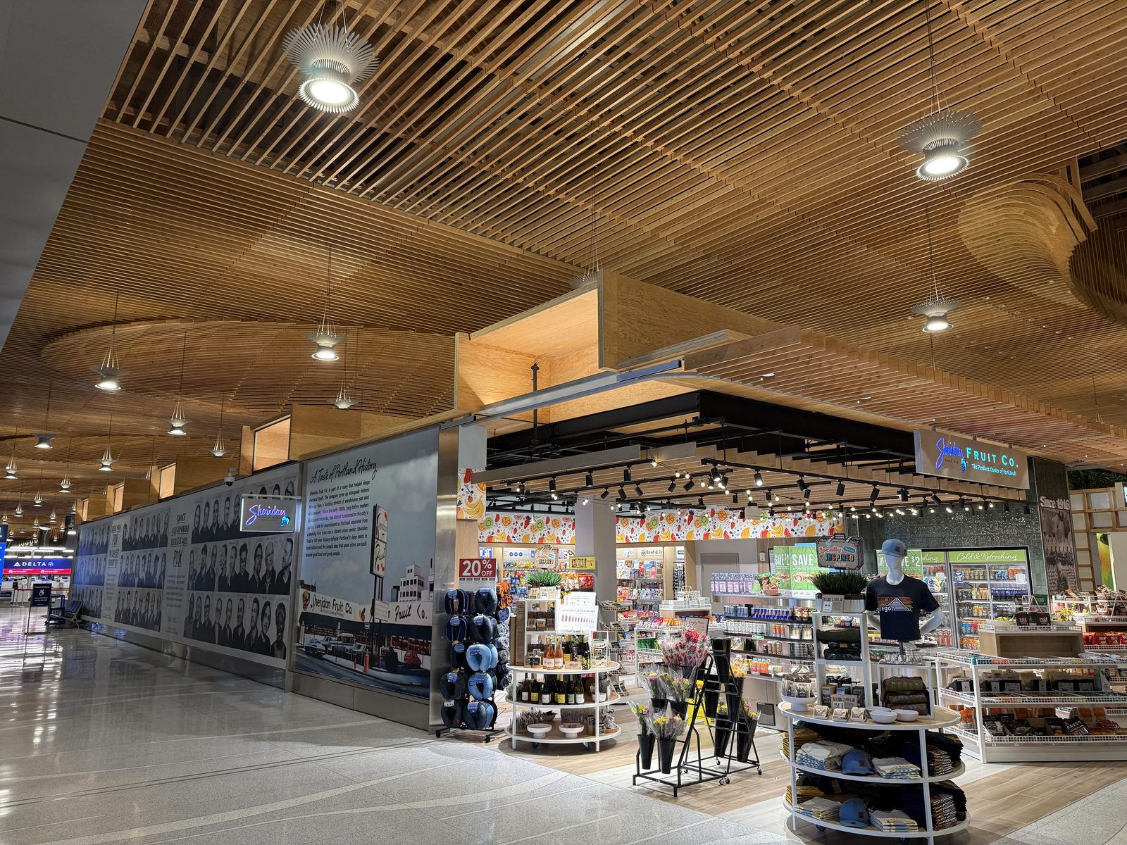 The Sheridan Fruit Co. storefront inside Portland International Airport, a lit sign reading Sheridan Fruit Co, The Produce Center of Portland, beside a black-and-white mural titled A Taste of Portland History, all under the terminal's curved wood-slat ceiling