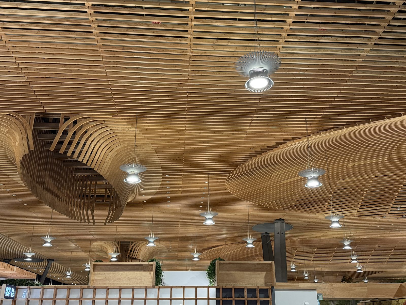 A close-up, upward view of Portland International Airport's undulating wood-slat ceiling at night, curved timber ribs sweeping overhead in a wave pattern, hung with pendant lights shaped like open sunbursts, seen from the terminal floor below