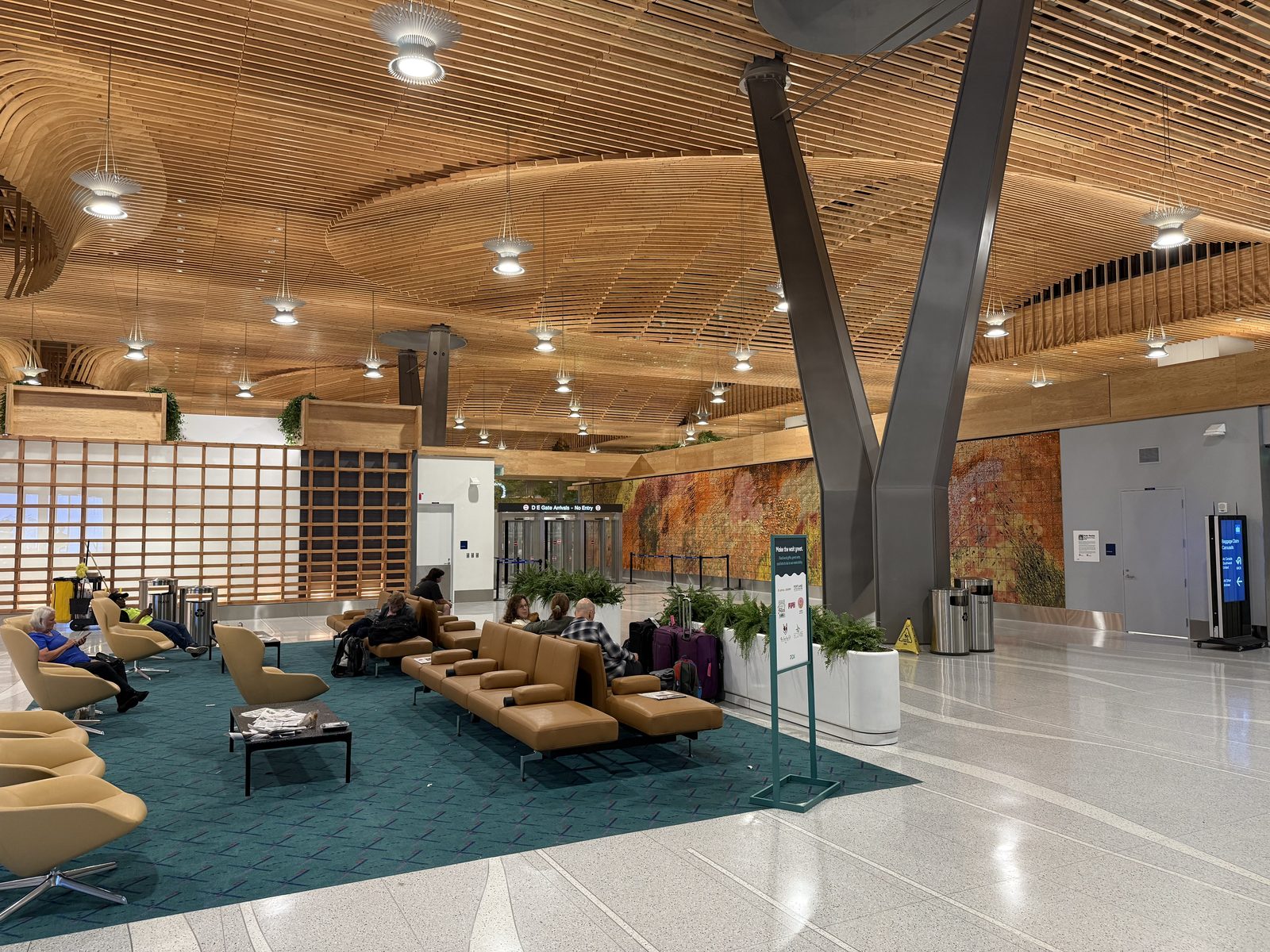 A wide interior view of Portland International Airport's timber-ceilinged terminal at night, showing curved wood-slat ceiling panels over a baggage claim seating area with tan leather chairs on a teal patterned carpet, travelers seated among their luggage, and a large abstract orange and red mural on the far wall