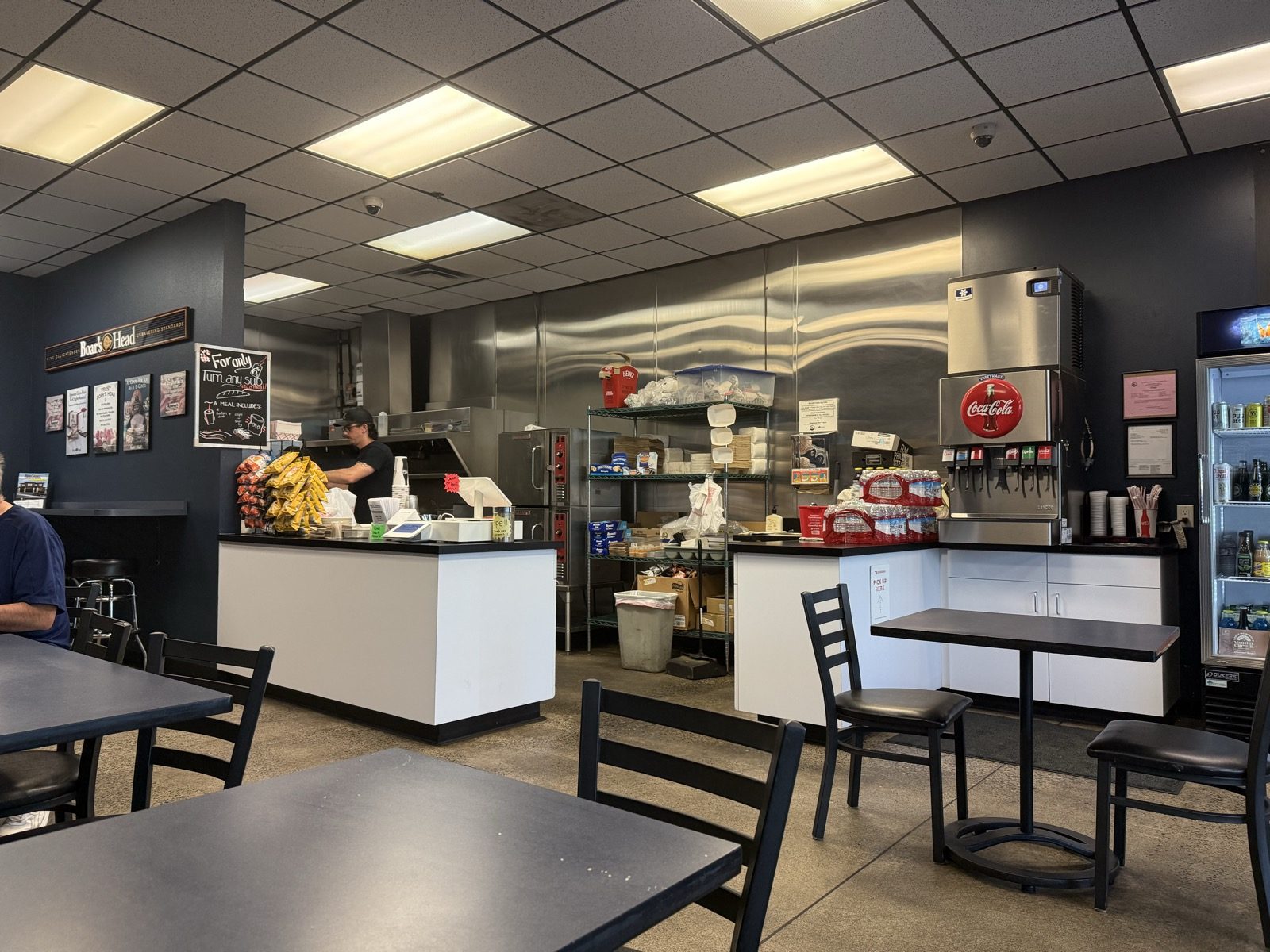 The order counter at Philly Bilmos with a rack of chips, a stainless steel kitchen behind it, a Coca-Cola fountain machine, a cooler of bottled drinks and Boar's Head signage on the wall