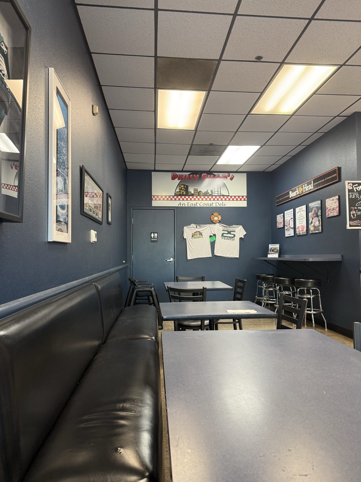 The dining room at Philly Bilmos, navy blue walls with a black vinyl banquette, dark tables and bar stools, under a sign reading Philly Bilmos, An East Coast Deli, with two t-shirts pinned to the wall, one of them celebrating 25 years