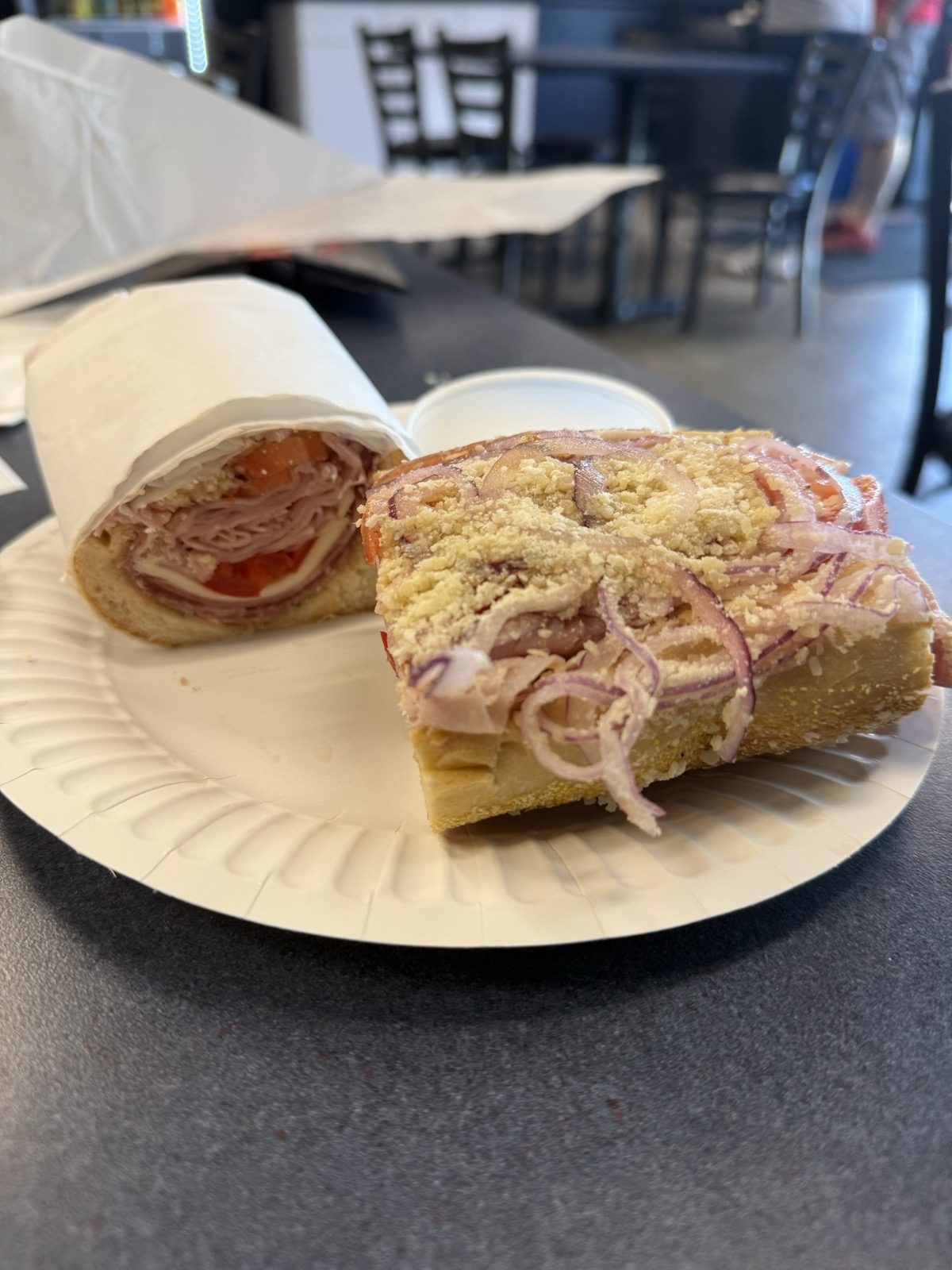 An Italian sub from Philly Bilmos in Vancouver, Washington, cut in half on a paper plate, one half still wrapped in white deli paper and the other laid open face-up showing ham, capicola and salami under tomato, rings of red onion and a heavy dusting of grated parmesan