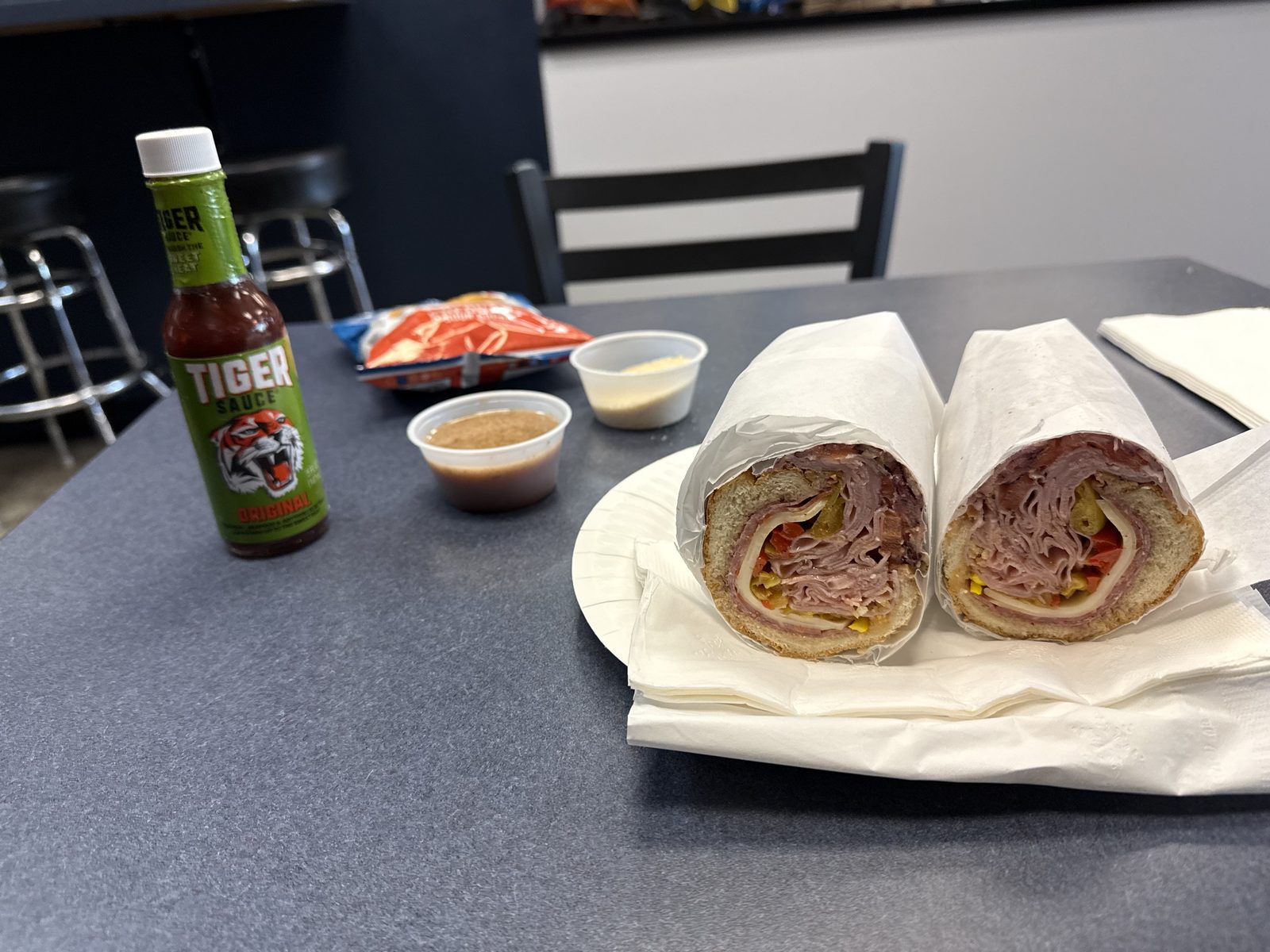 An Italian sub from Philly Bilmos cut in half and wrapped in white deli paper on a paper plate, showing a cross-section of shaved ham, capicola and salami rolled around provolone, pepperoncini and red onion, next to a bottle of Tiger Sauce, a small cup of oil-and-vinegar dressing, a small cup of grated parmesan, and a bag of chips on a black table