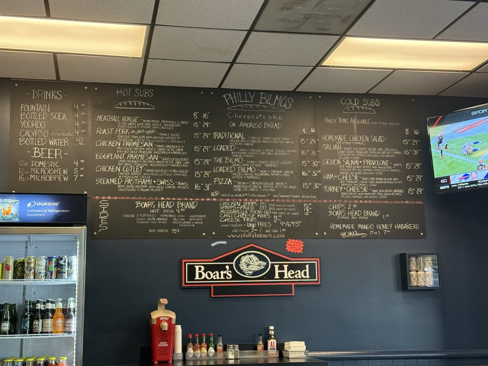 The chalkboard menu at Philly Bilmos in Vancouver, Washington, with hand-lettered columns for Drinks, Hot Subs, Philly Bilmos Cheesesteaks on Amoroso Bread, and Cold Subs, above a Boar's Head sign and a counter of condiments
