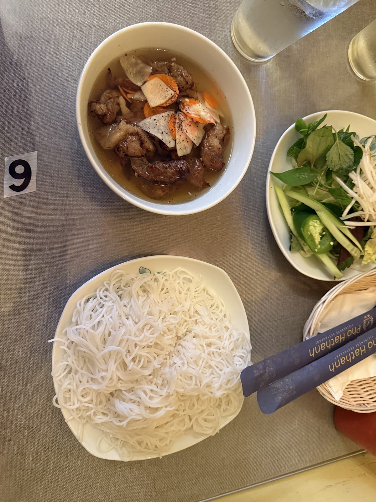 A bowl of bún chả Hanoi at Pho Hathanh in Portland, Oregon — grilled pork and pork belly in a sweet-savory broth with pickled carrot and daikon, next to a plate of rice vermicelli and a plate of fresh herbs, with a chopstick sleeve printed with the restaurant's name