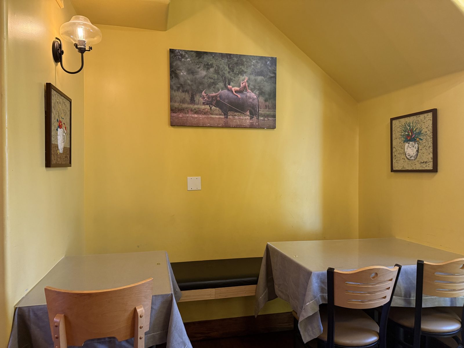 A small mustard-yellow dining nook at Pho Hathanh with two linen-covered tables and a photograph of a boy asleep on the back of a water buffalo hanging on the wall