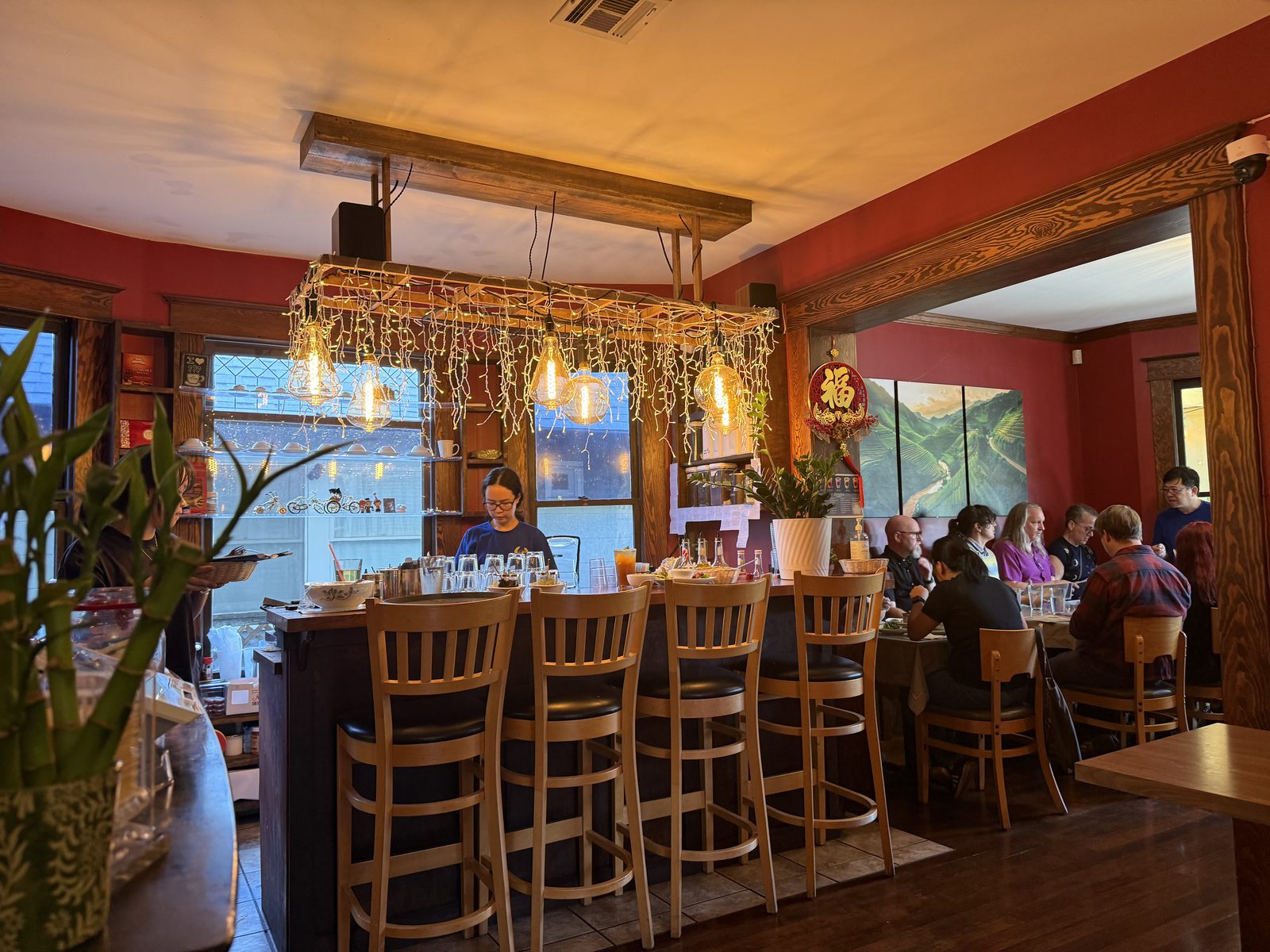 The bar at Pho Hathanh — wooden stools under a beam hung with string lights and Edison bulbs, a red accent wall with a mural of terraced rice paddies, and diners seated at tables beyond it