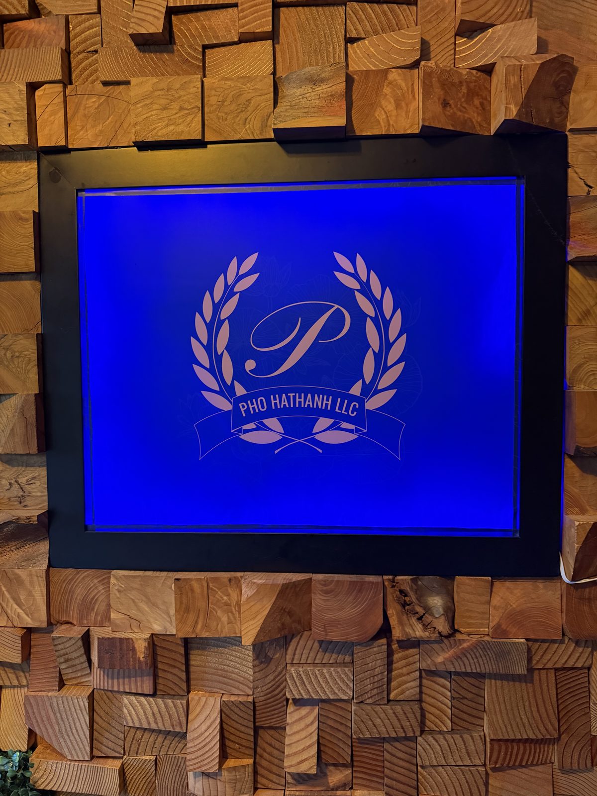 A backlit blue Pho Hathanh LLC logo — a laurel wreath around a script letter P — set into a wall of stacked reclaimed wood blocks just inside the entrance