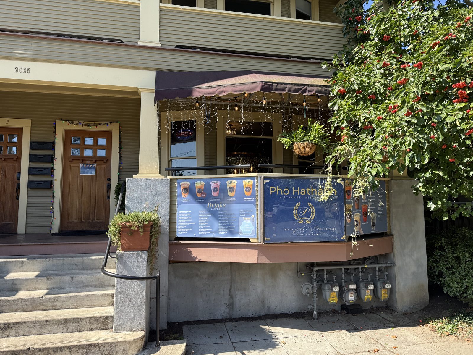 The Pho Hathanh storefront in Portland, Oregon — a converted craftsman house with a drinks-menu sign board, a lit OPEN sign, string lights under the porch awning, and a flowering vine climbing the porch post