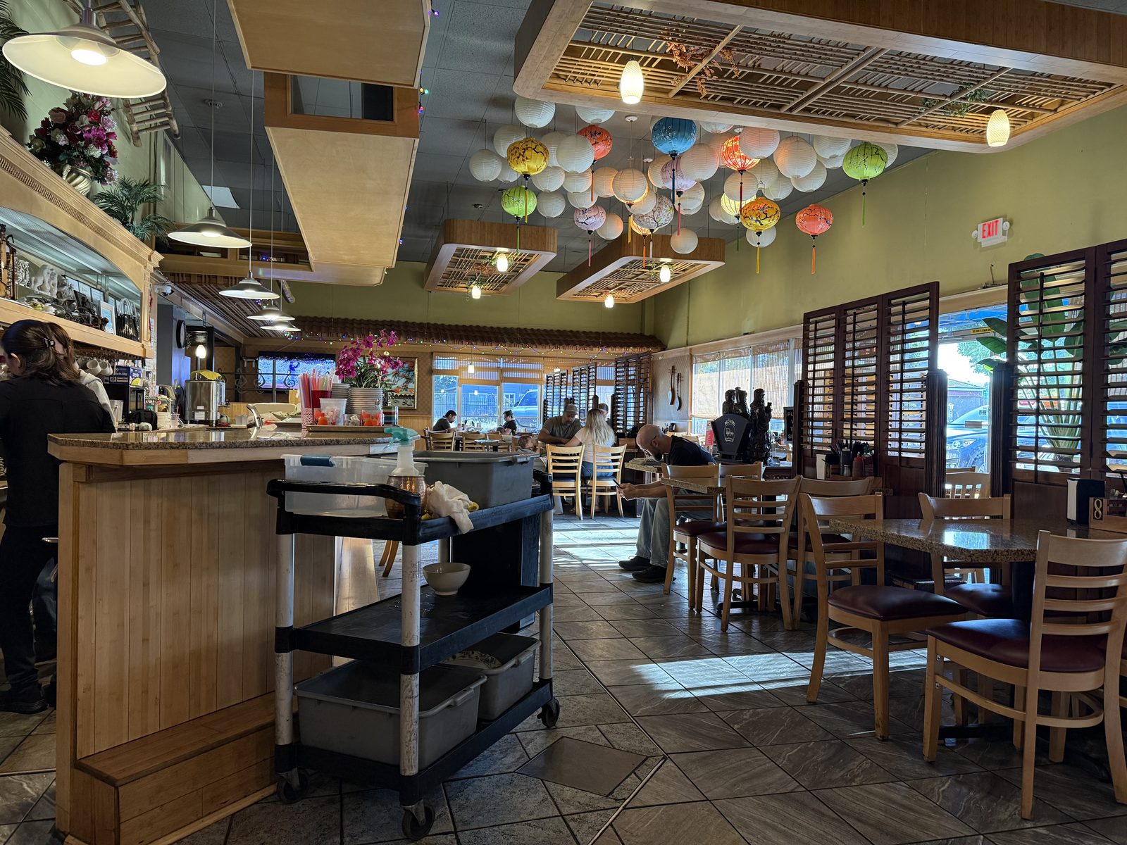 The dining room at Pho Hung on 82nd Avenue in Portland, Oregon — a ceiling hung with colorful paper lanterns, sage-green walls, wood-shuttered windows, and tile floors, with customers seated at wooden tables