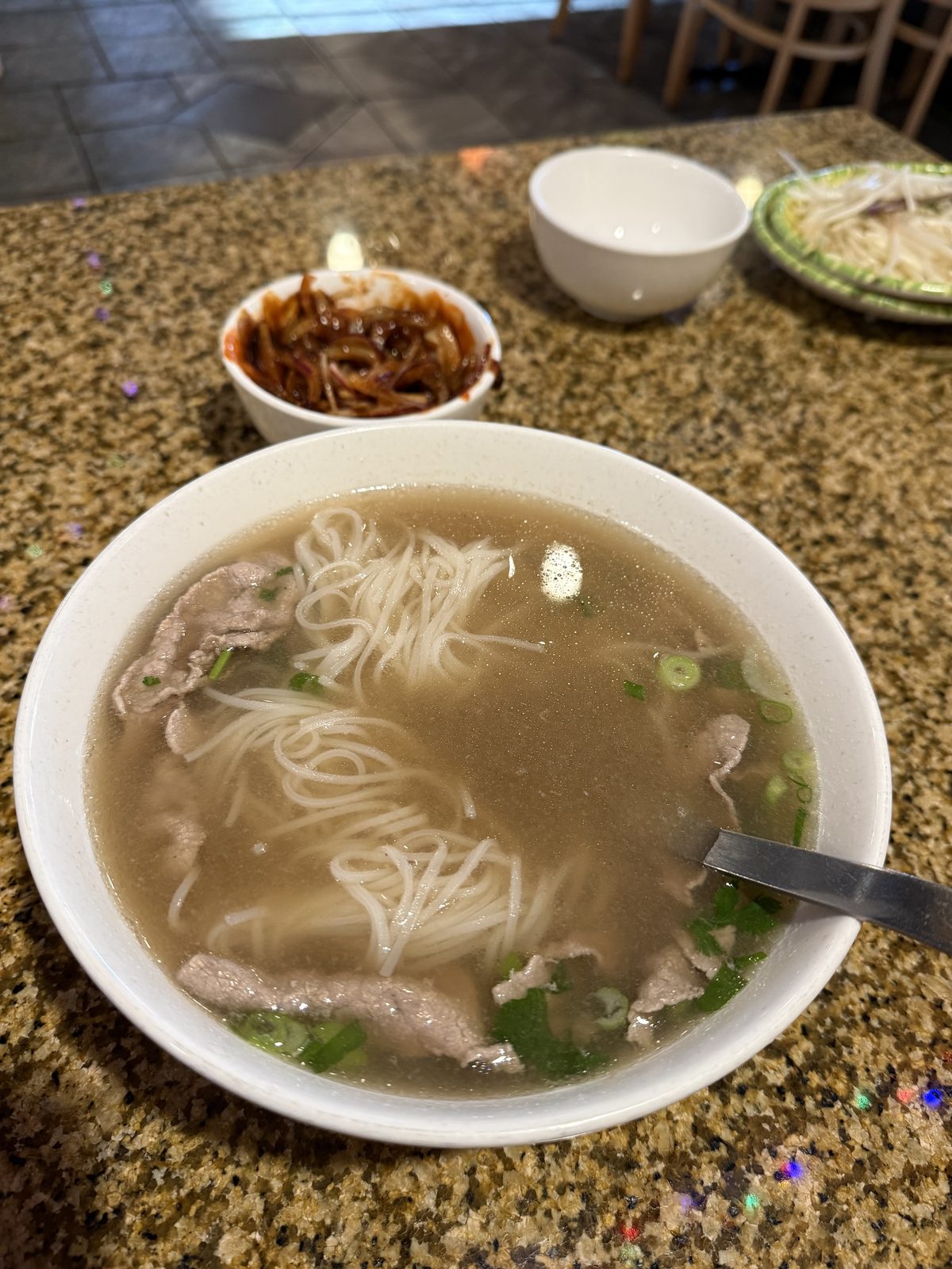 A steaming bowl of beef pho at Pho Hung on 82nd in Portland, Oregon — rice noodles and thin-sliced beef in broth topped with scallions and cilantro, a spoon resting in the bowl, with two small cups alongside it on the counter