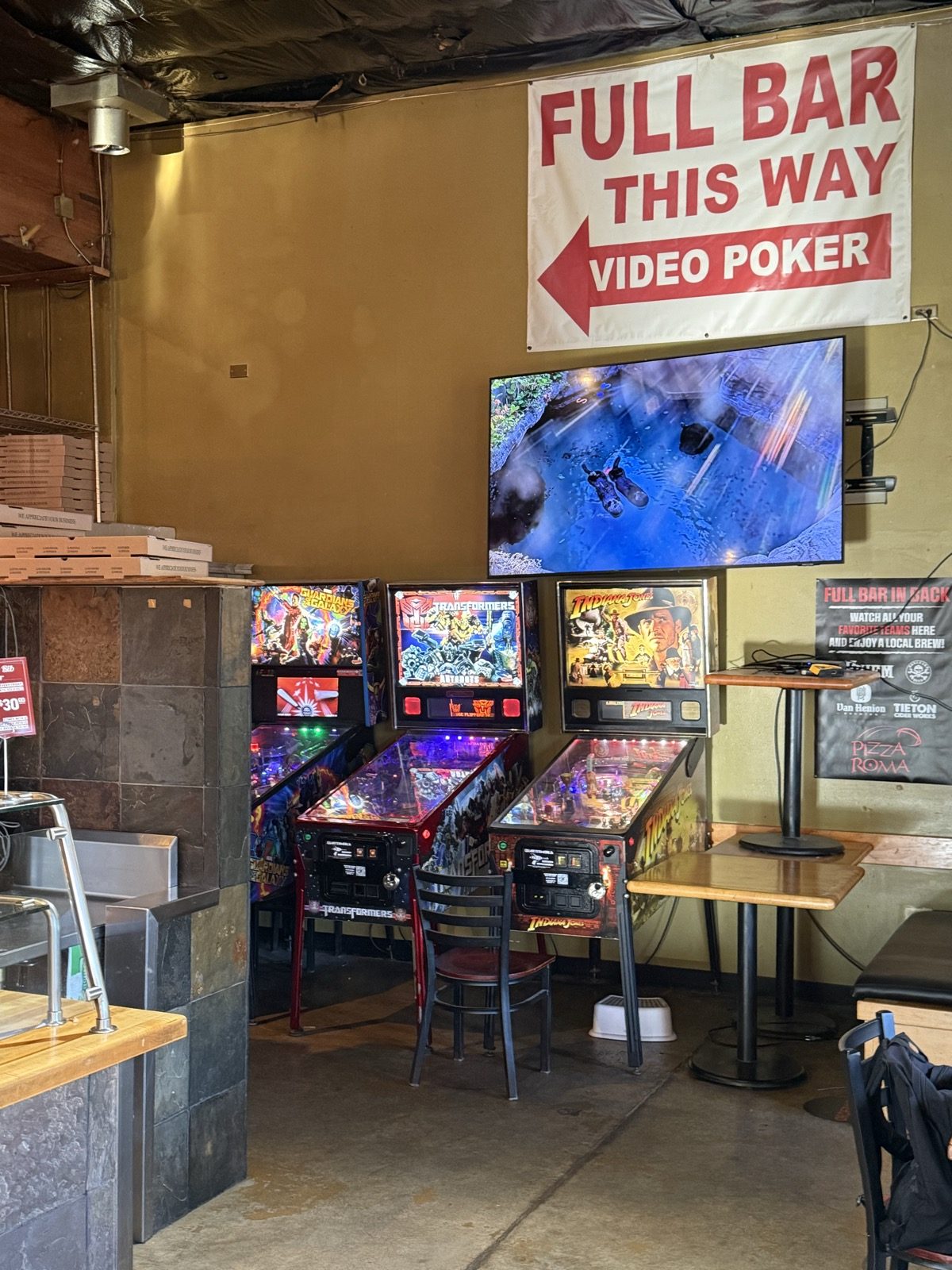 A row of pinball machines under a mounted TV at Pizza Roma in Portland, Oregon, with a hand-lettered sign reading Full Bar This Way, Video Poker pointing toward the back room