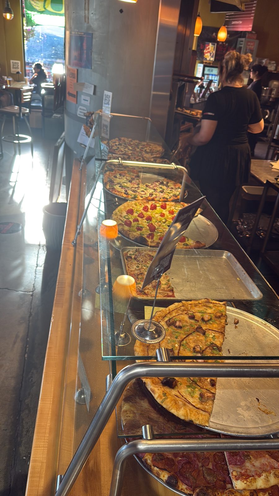 The pizza case at Pizza Roma in Portland, Oregon, several whole pies under glass with hand-written chalkboard labels, a staff member visible behind the counter
