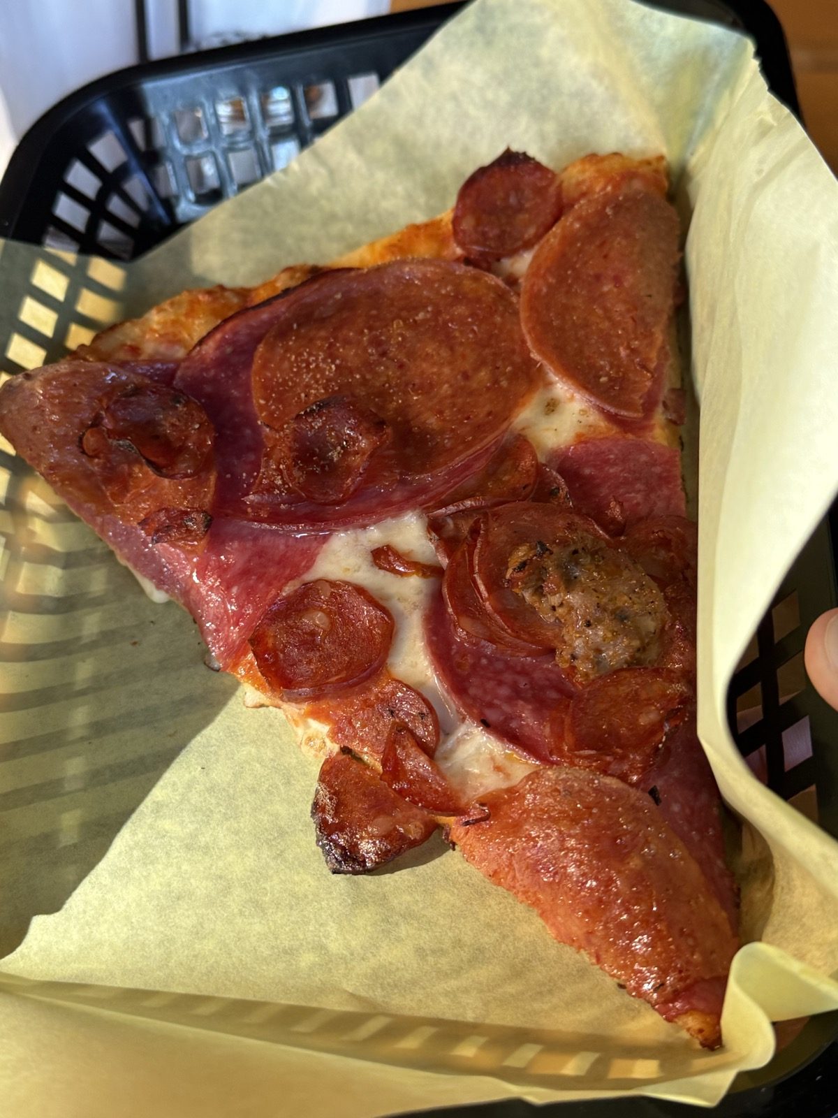 A slice of meat-lover's pizza topped with pepperoni, salami and sausage, held in a black plastic food basket lined with parchment paper, from Pizza Roma in the Woodstock neighborhood of Portland, Oregon