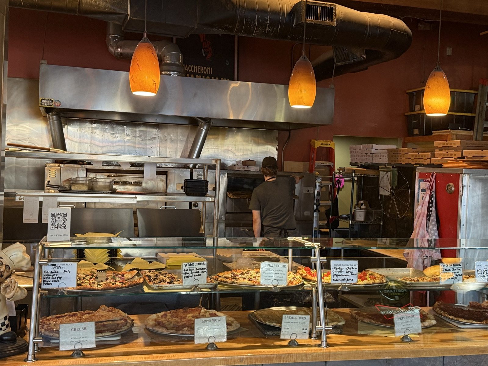 A cook working at the pizza oven behind the counter at Pizza Roma, with the pizza case in the foreground holding cheese, pepperoni and breadsticks
