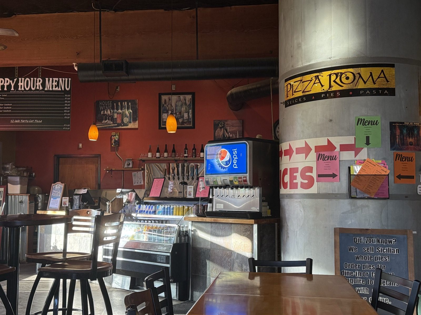 The Pizza Roma logo on a concrete support column reading Pizza Roma, Slices and Pies, Pasta, next to a happy hour menu chalkboard, a beer cooler and a self-serve soda fountain