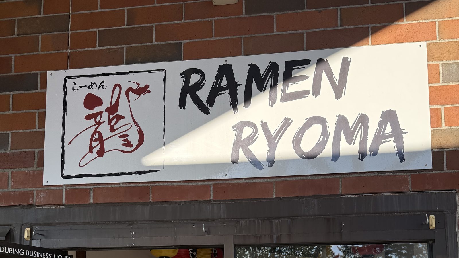The Ramen Ryoma sign on a brick wall — the shop's red dragon calligraphy logo in a black box, beside RAMEN RYOMA in brushed black letters