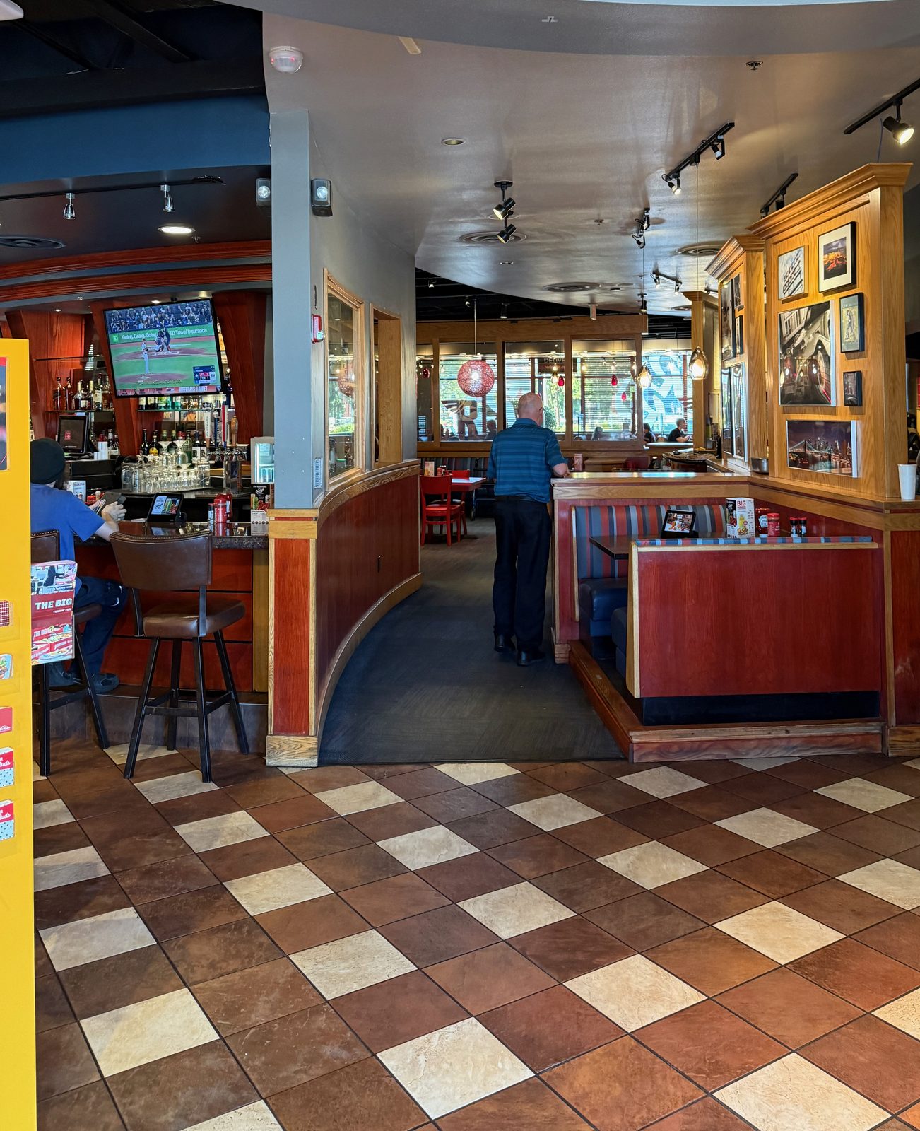 The interior of the Red Robin — a bar on the left with a baseball game on the television, red vinyl booths, warm wood trim, a wall of framed photographs, and a brown-and-cream checkerboard tile floor