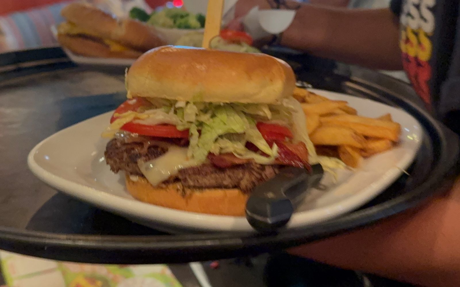 The MadLove burger arriving on a black tray — a thick beef patty with white melted cheese draped over the side, strips of red pepper relish, shredded lettuce and tomato under a glossy golden bun held together with a wooden pick, with a pile of steak fries beside it on a white plate