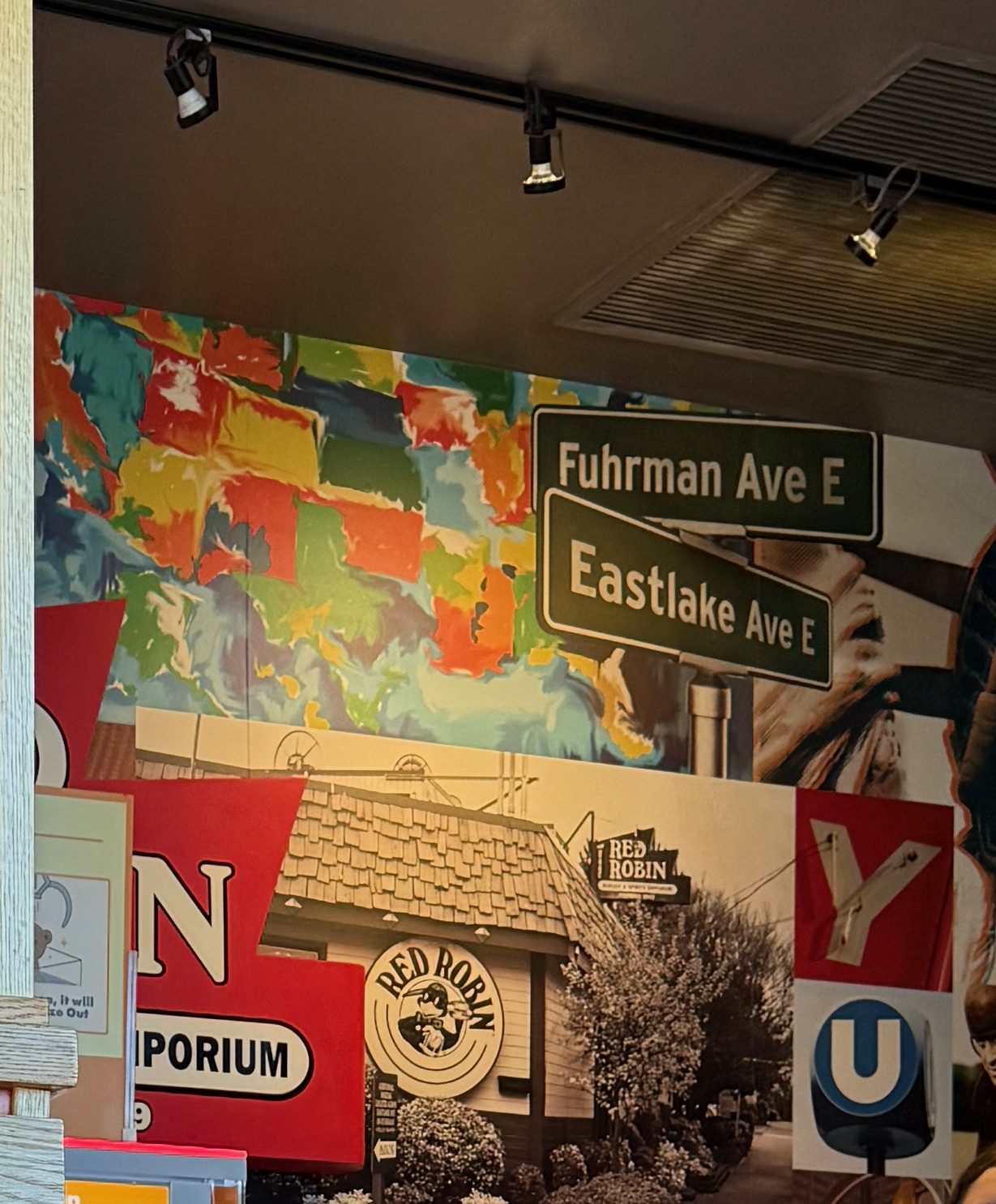 A collage mural in the Red Robin waiting area, showing two crossed green Seattle street signs reading Fuhrman Ave E and Eastlake Ave E above a sepia photograph of a small shingled corner building with a circular Red Robin logo sign and a sign reading Red Robin Burger and Spirits Emporium