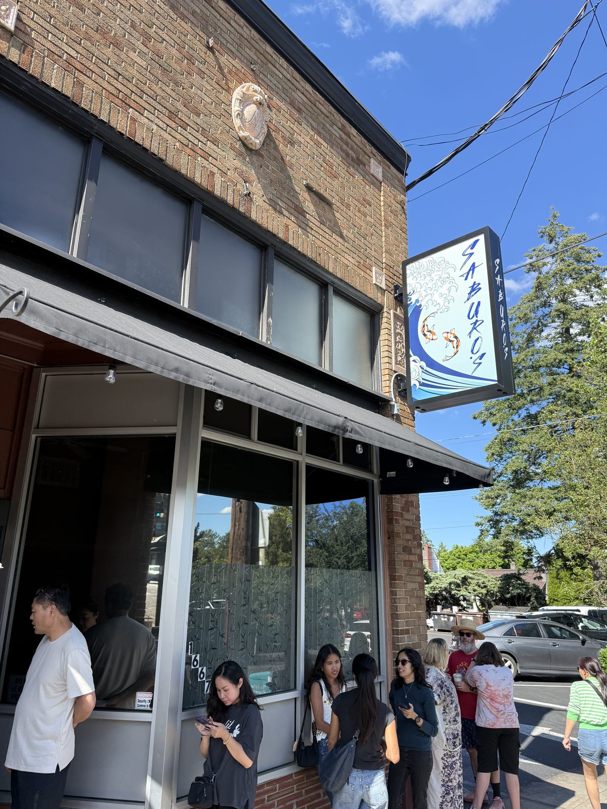 The brick storefront of Saburo's Sushi House in the Sellwood neighborhood of Portland, Oregon, with a hanging blue sign painted with a wave and two koi fish reading SABURO'S, a black awning over street-level windows marked 1667, and a small crowd of customers waiting on the sidewalk out front in late-afternoon sun