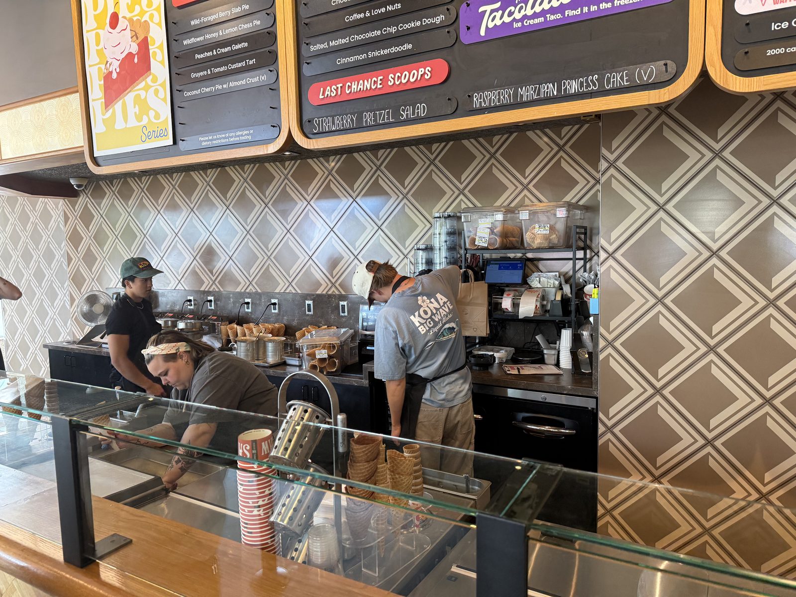 A scoop counter at Salt & Straw with a case of ice cream tubs, stacked waffle cones, and a chalkboard menu overhead advertising the Summer Pies ice cream series