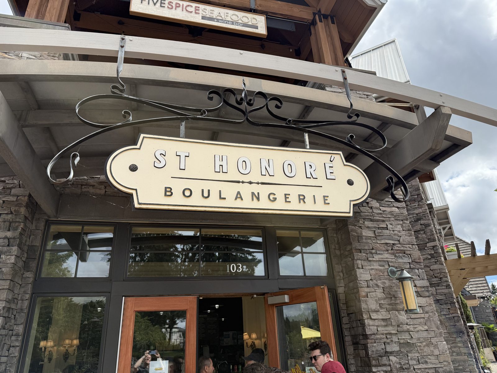 The cream-and-black hanging sign for St Honoré Boulangerie above a stone storefront, with yellow café umbrellas and a busy outdoor patio on the sidewalk below