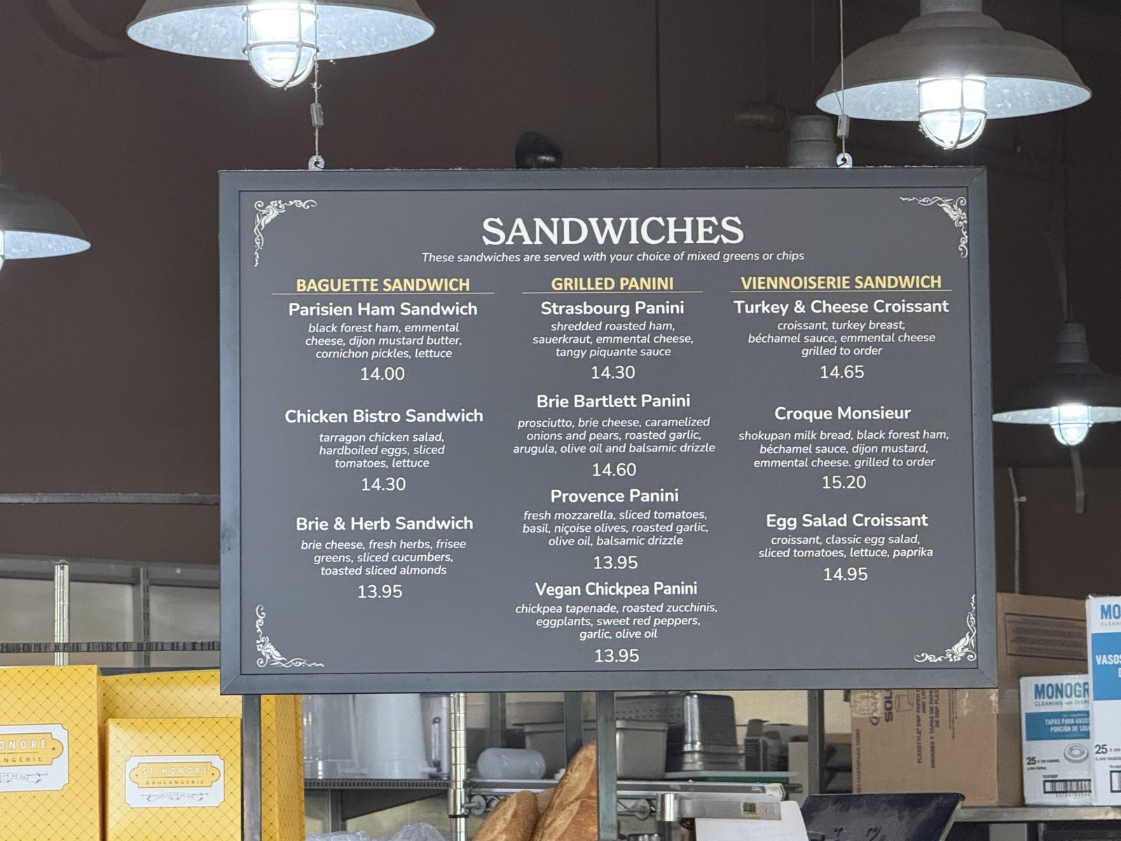 A hanging menu board at St Honoré Boulangerie listing Sandwiches in three columns — Baguette Sandwich, Grilled Panini and Viennoiserie Sandwich — with prices from $13.95 to $15.20