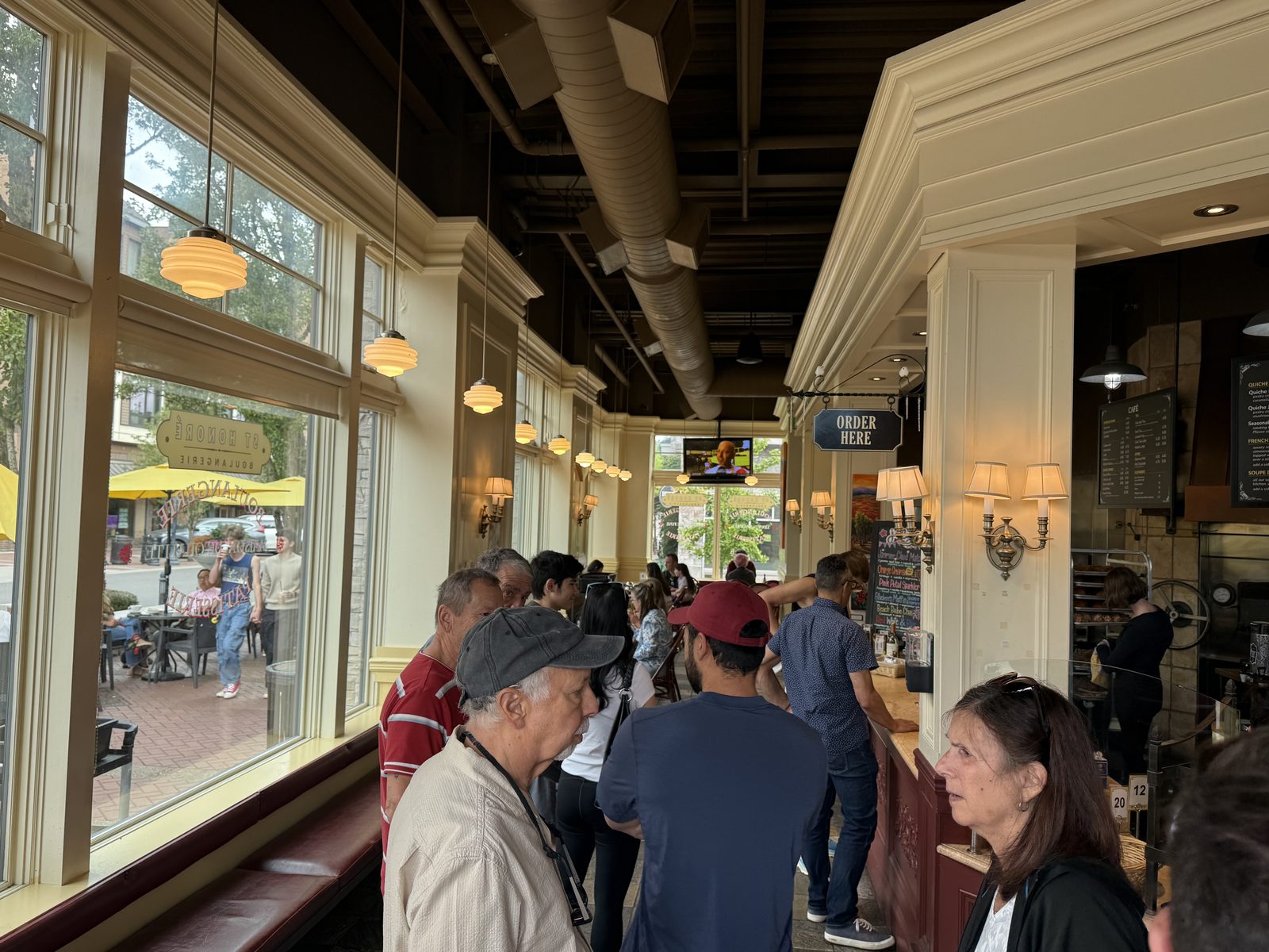 A line of customers waiting to order inside St Honoré Boulangerie, under paper lanterns and a large window looking out onto First Street, with an Order Here sign overhead