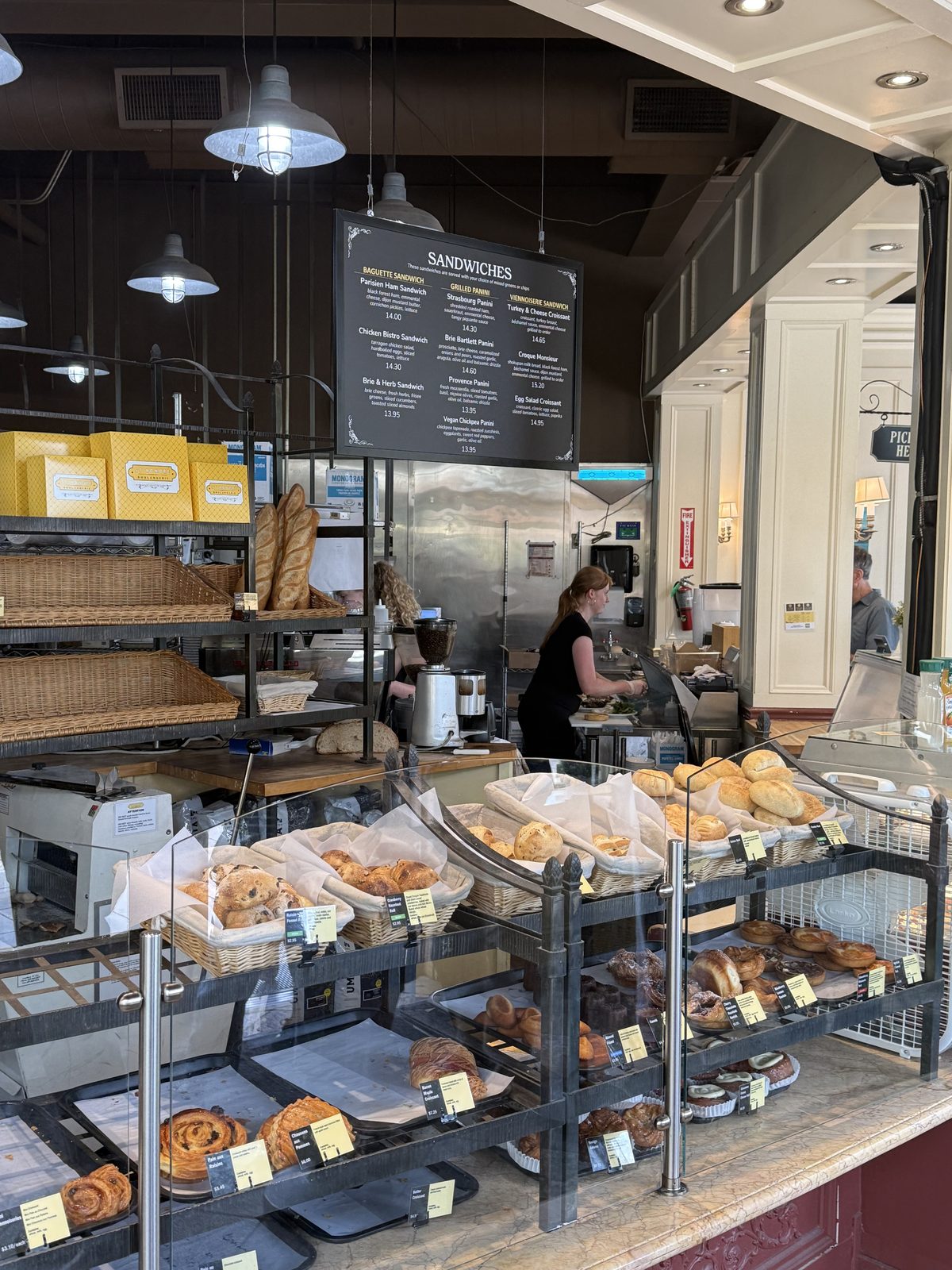 A glass bakery case filled with baskets of baguettes and rolls, croissants, chocolate pastries and cookies, with a hand-lettered sandwich menu board hanging above
